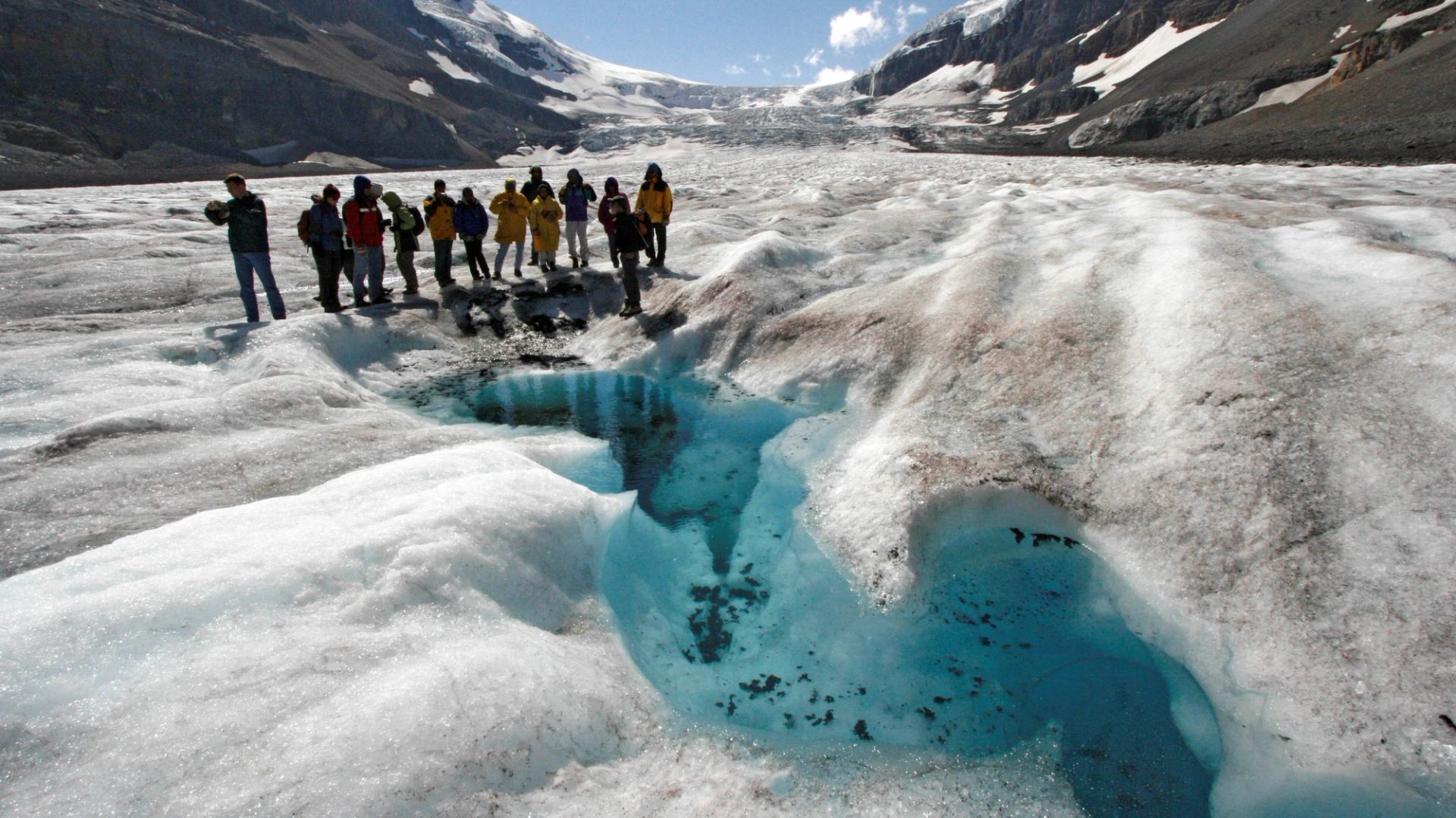 Group stands on glacier beside blue meltwater pool, mountain peaks in background.