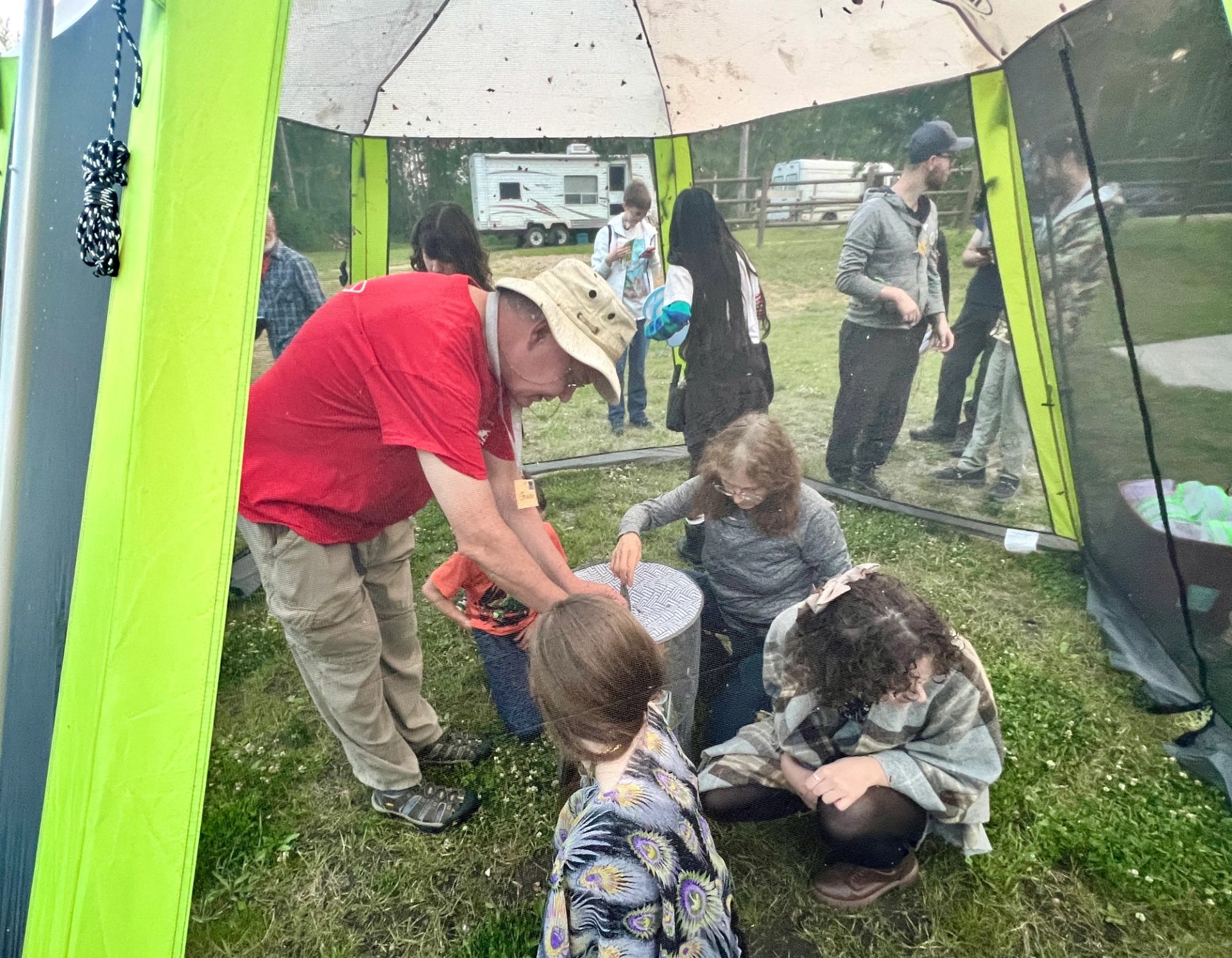 People gather inside a mesh tent, some kneeling around a small round table.