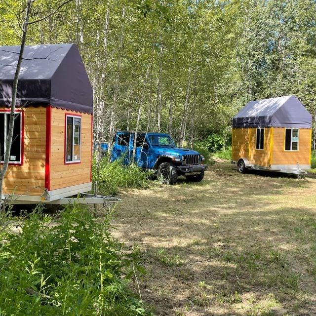 Two tiny cabins and a blue SUV parked in a sunny forest clearing.