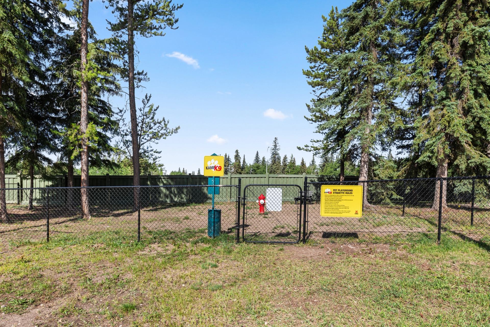 A fenced dog park features open space, pet waste stations, and tall trees under a blue sky.