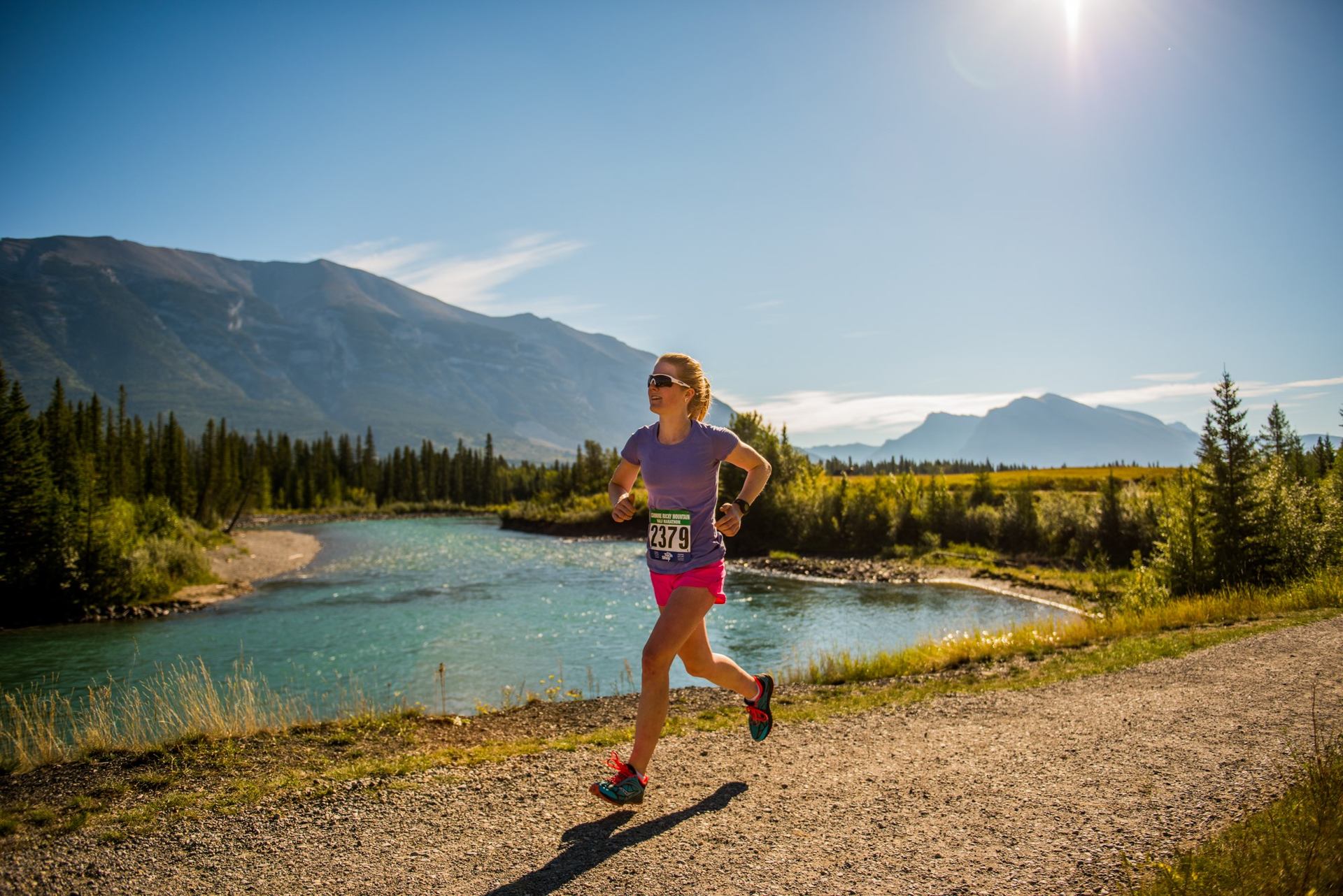 Runner with bib number 2379 along riverbank, Rocky Mountains and pine trees in background.