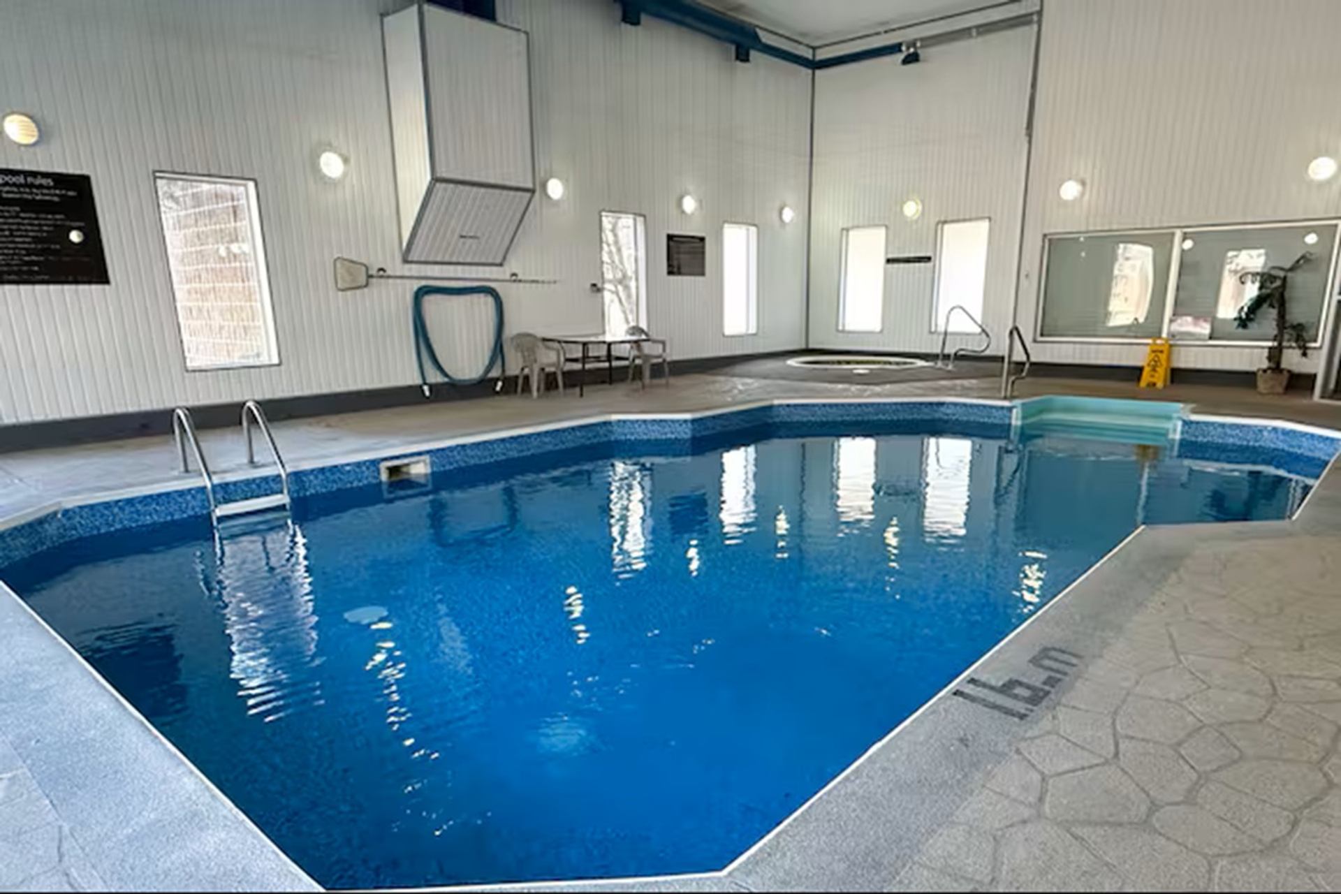 Indoor swimming pool with ladders, hot tub, chairs and safety sign on deck.