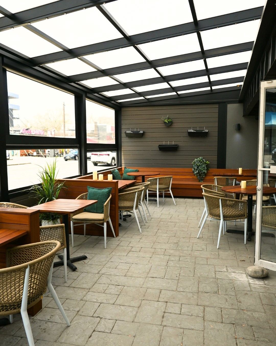 Patio seating area with wooden tables and chairs under a glass roof, restaurant entrance.