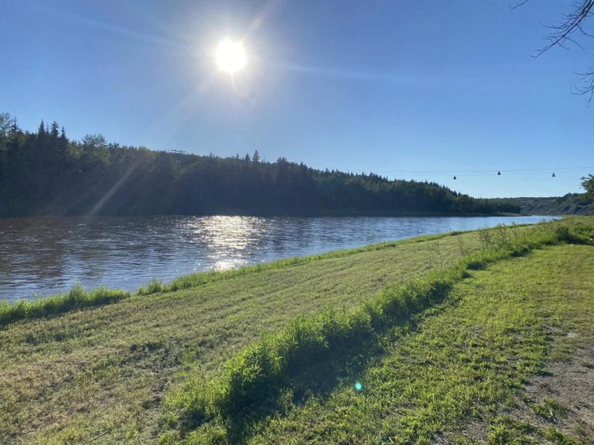 A grassy riverbank stretches alongside calm water under a bright sky at Tail Creek Park.
