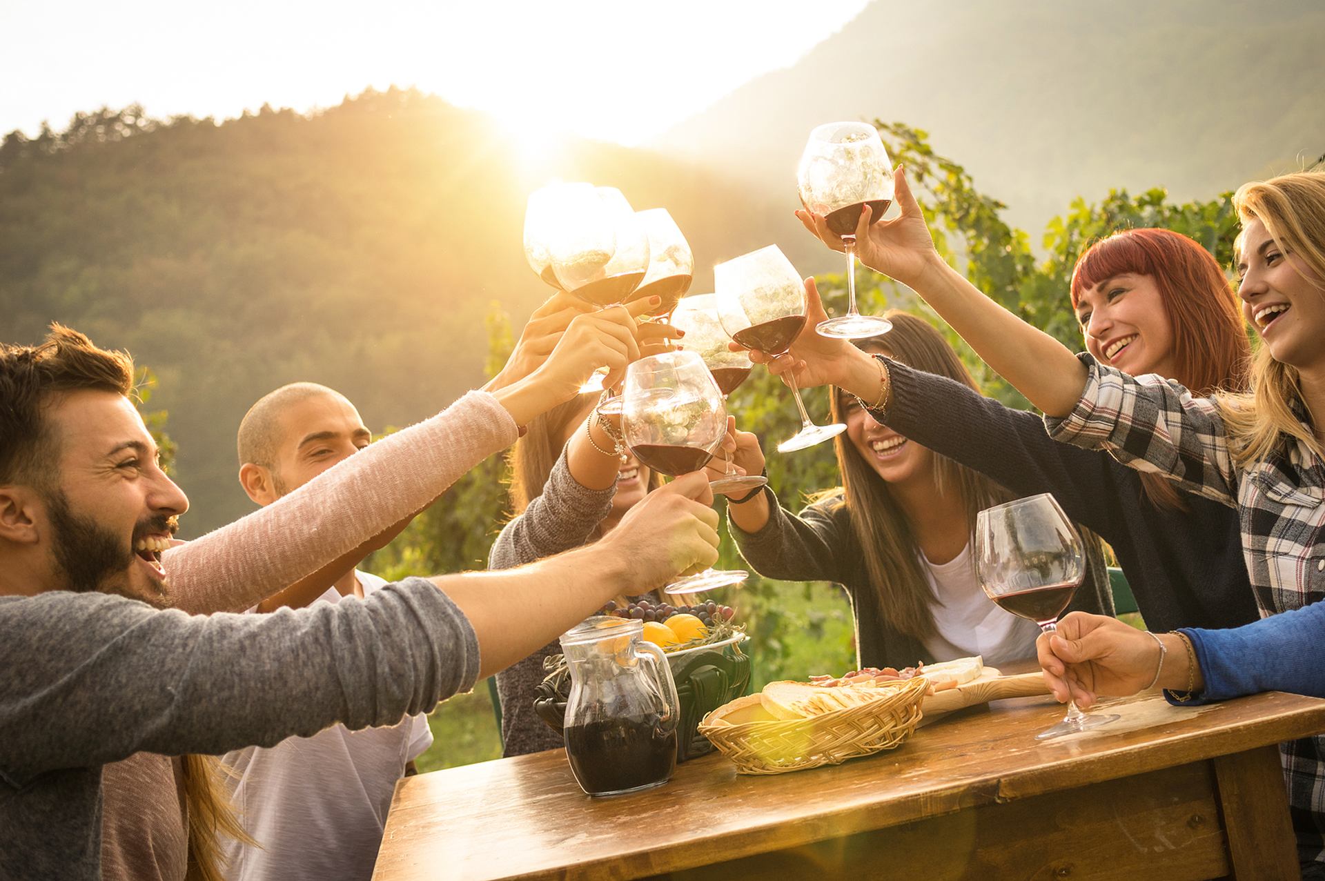 Friends toasting wine at sunset around a rustic outdoor table in a vineyard.