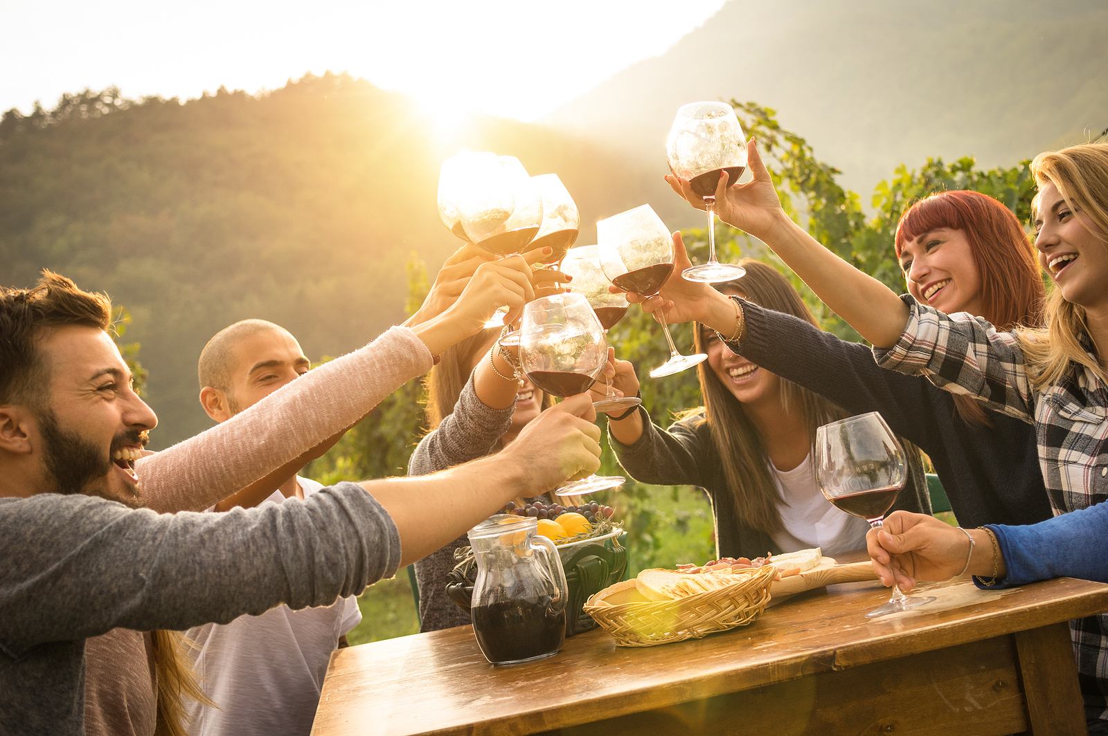Friends toasting wine at sunset around a rustic outdoor table in a vineyard.