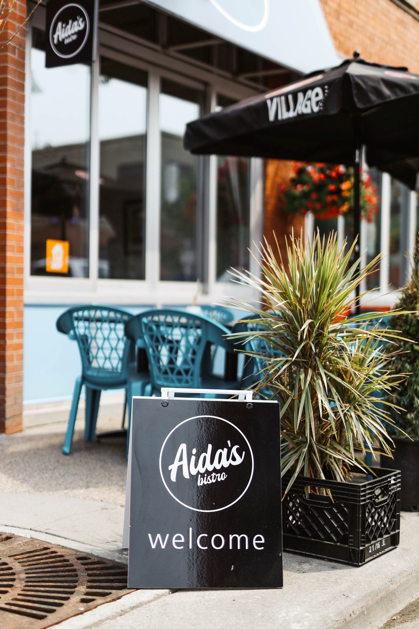 Outdoor café with a black “Aida’s welcome” sign and teal chairs near a brick wall.