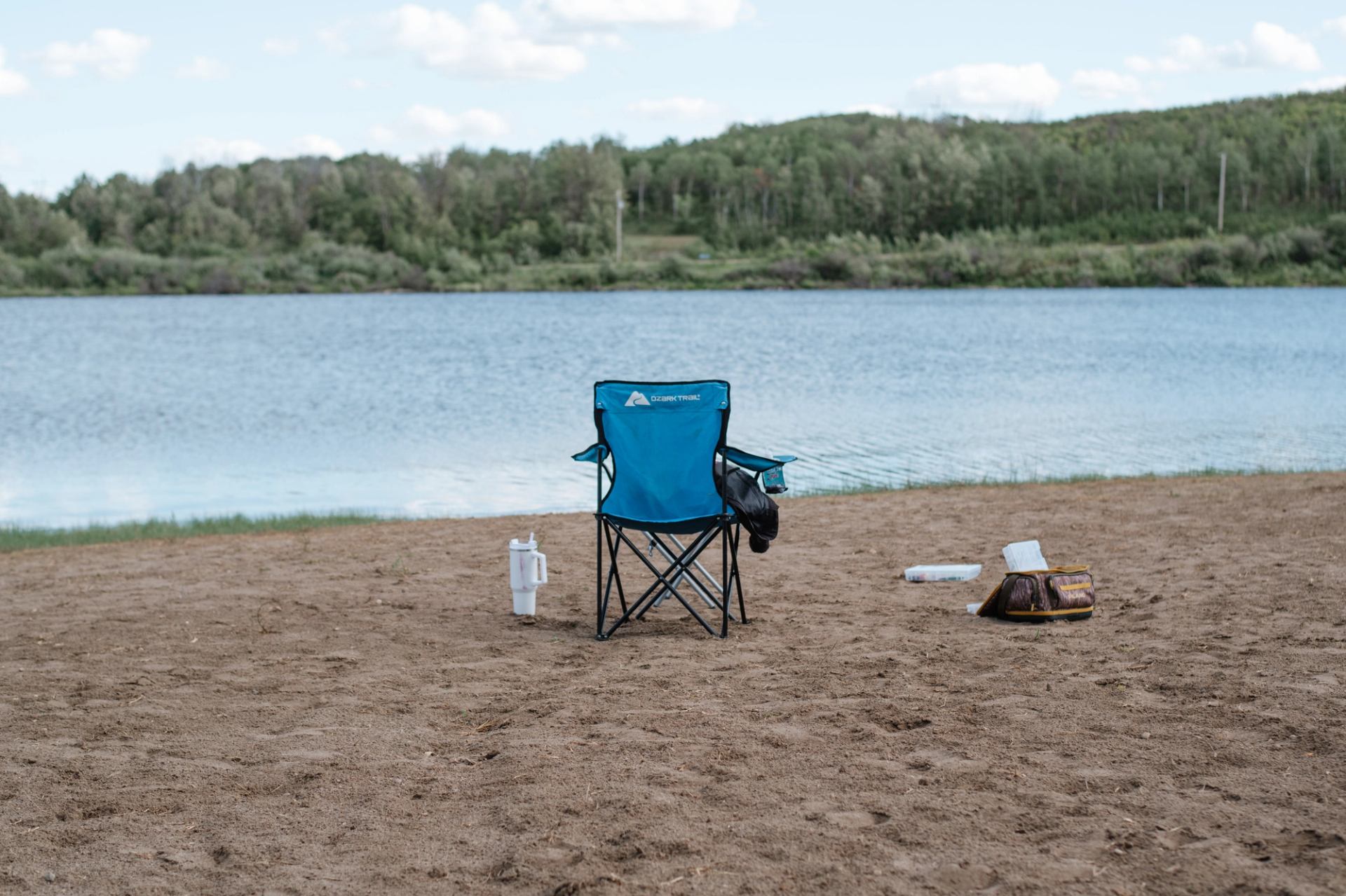 Blue camping chair on sandy beach facing calm lake with forested shoreline.
