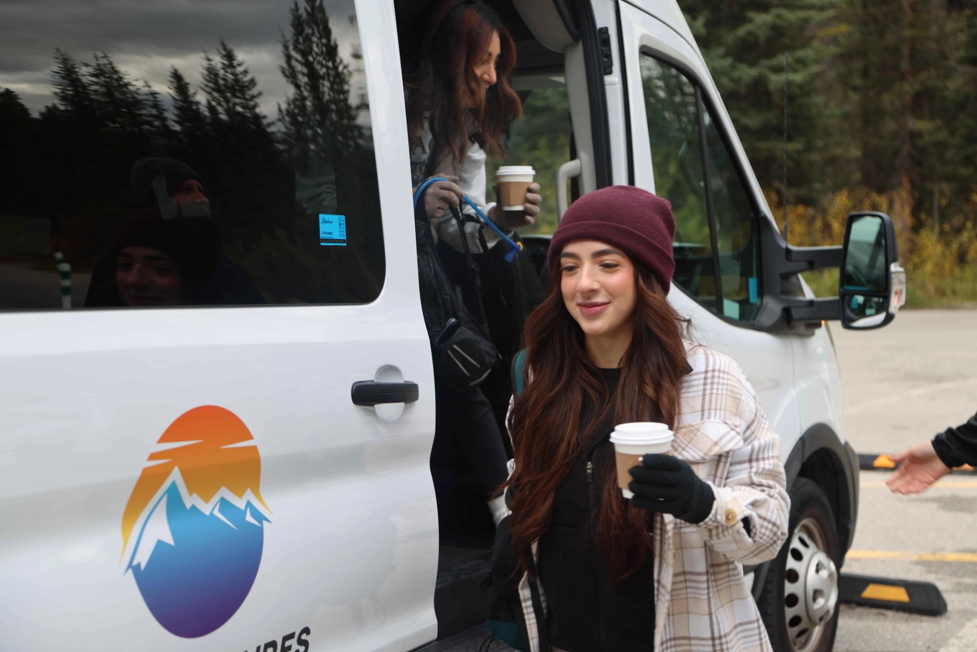 A woman in a beanie steps out of a white van with a mountain logo, holding a coffee cup.