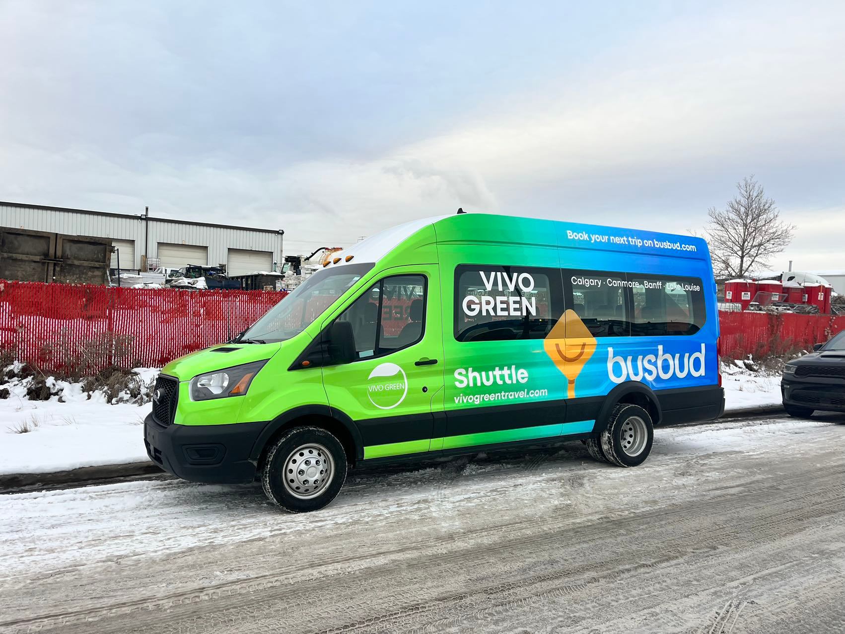 Green and blue Vivo Green Busbud shuttle van parked on a snow-covered road.