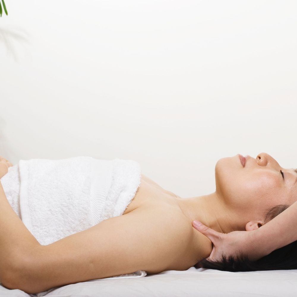 Woman lying on massage table with hands under neck, white towel and sheet visible.