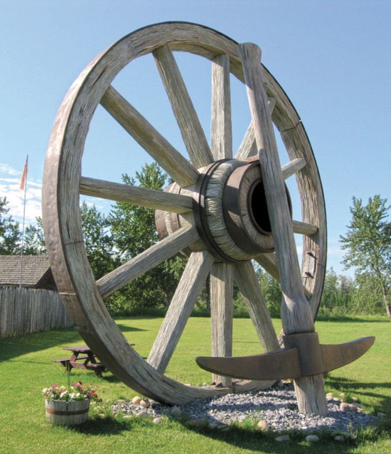 The image shows a giant wooden wagon wheel and pickaxe monument on a grassy lawn.