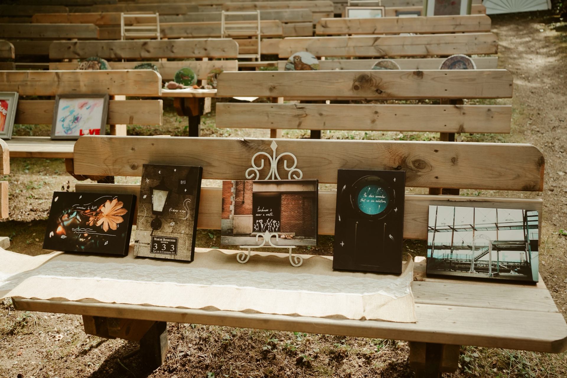 Framed artwork arranged on wooden benches in an outdoor exhibit area.