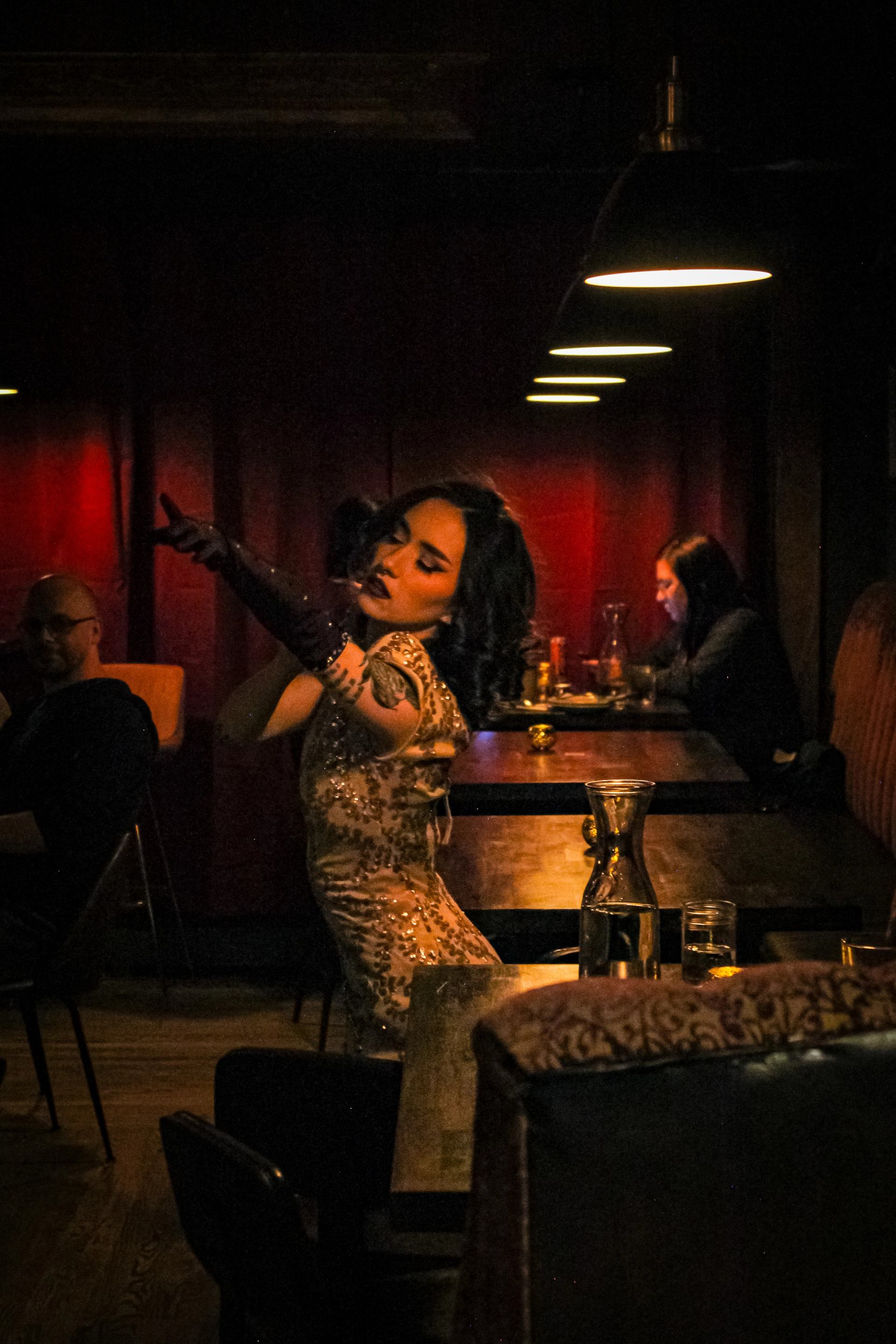 A woman in a sparkling dress and long gloves strikes a pose in a dimly lit room with red curtains.