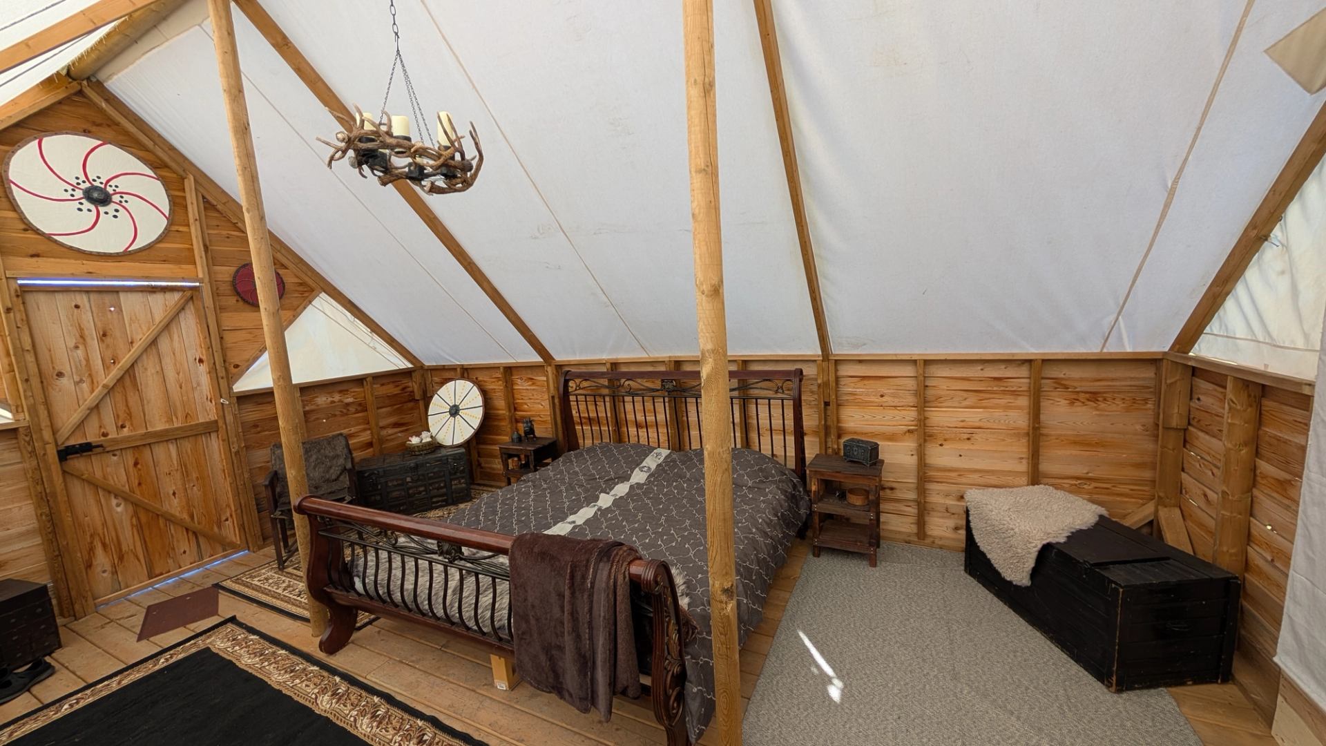 Rustic glamping tent bedroom with wooden walls, a bed, and an antler chandelier.