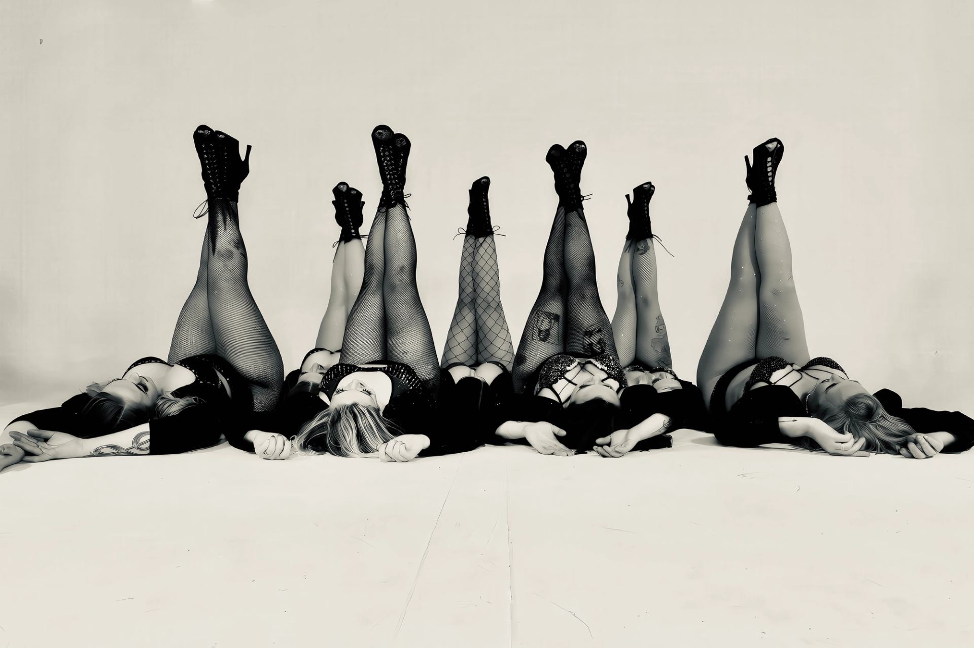 Black and white photo of several women lying on their backs with their legs raised, wearing fishnet stockings and high-heeled boots.