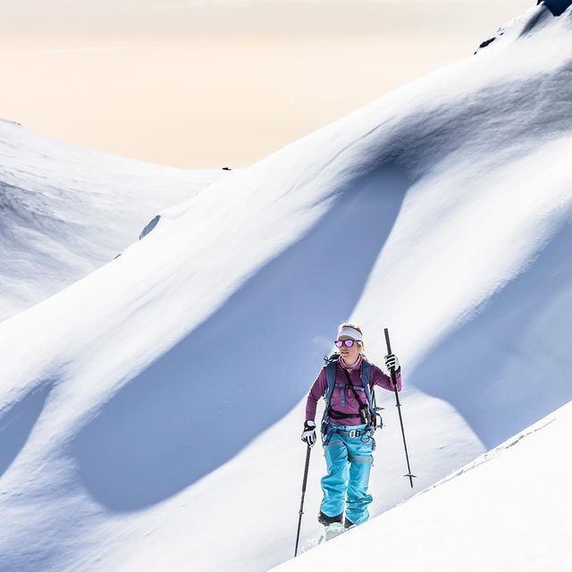 Skier on snow-covered mountain with poles, helmet, and backpack under clear skies.