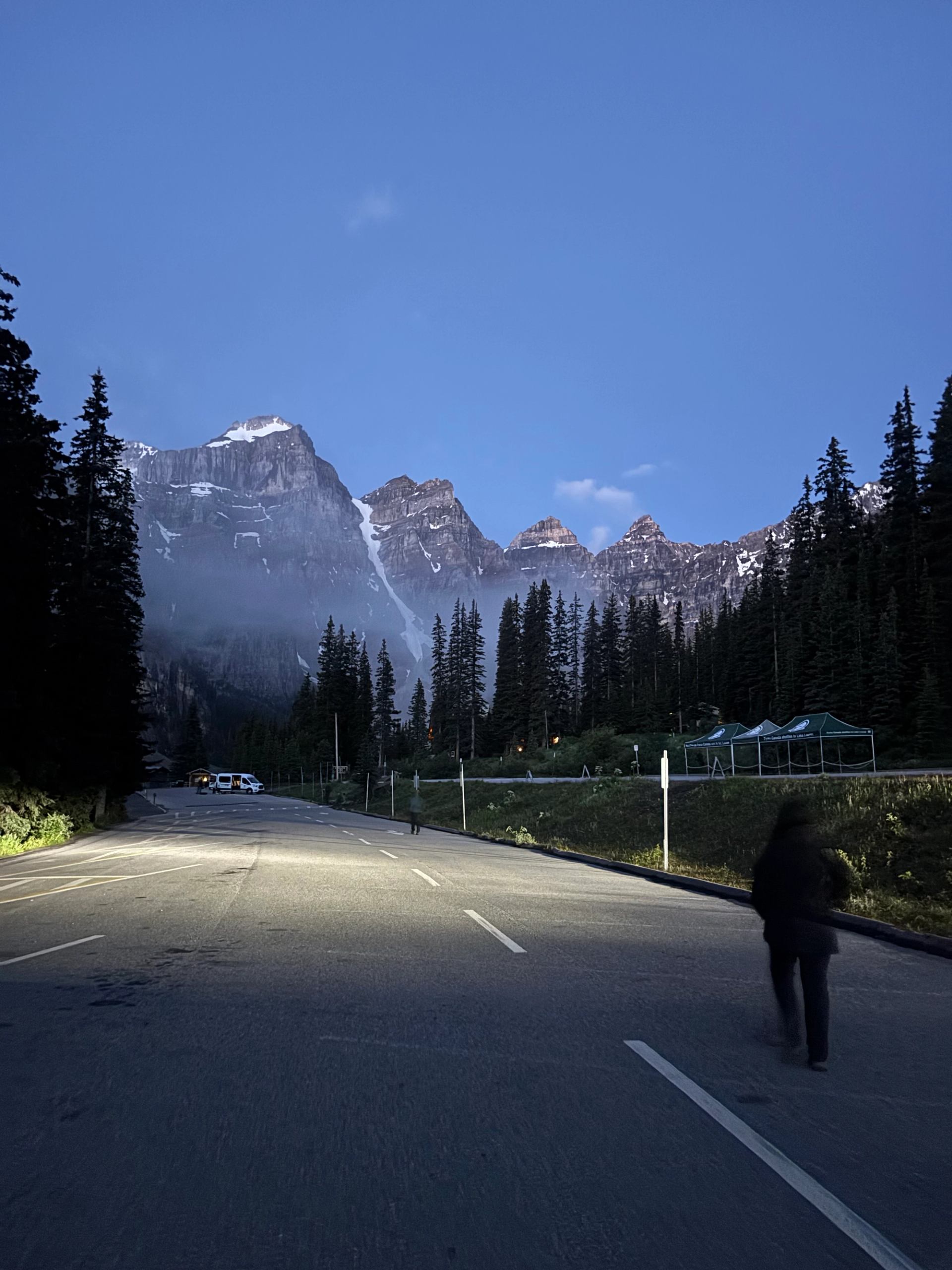 Mountain road with headlights illuminating pavement, person walking, and snowy peaks ahead.