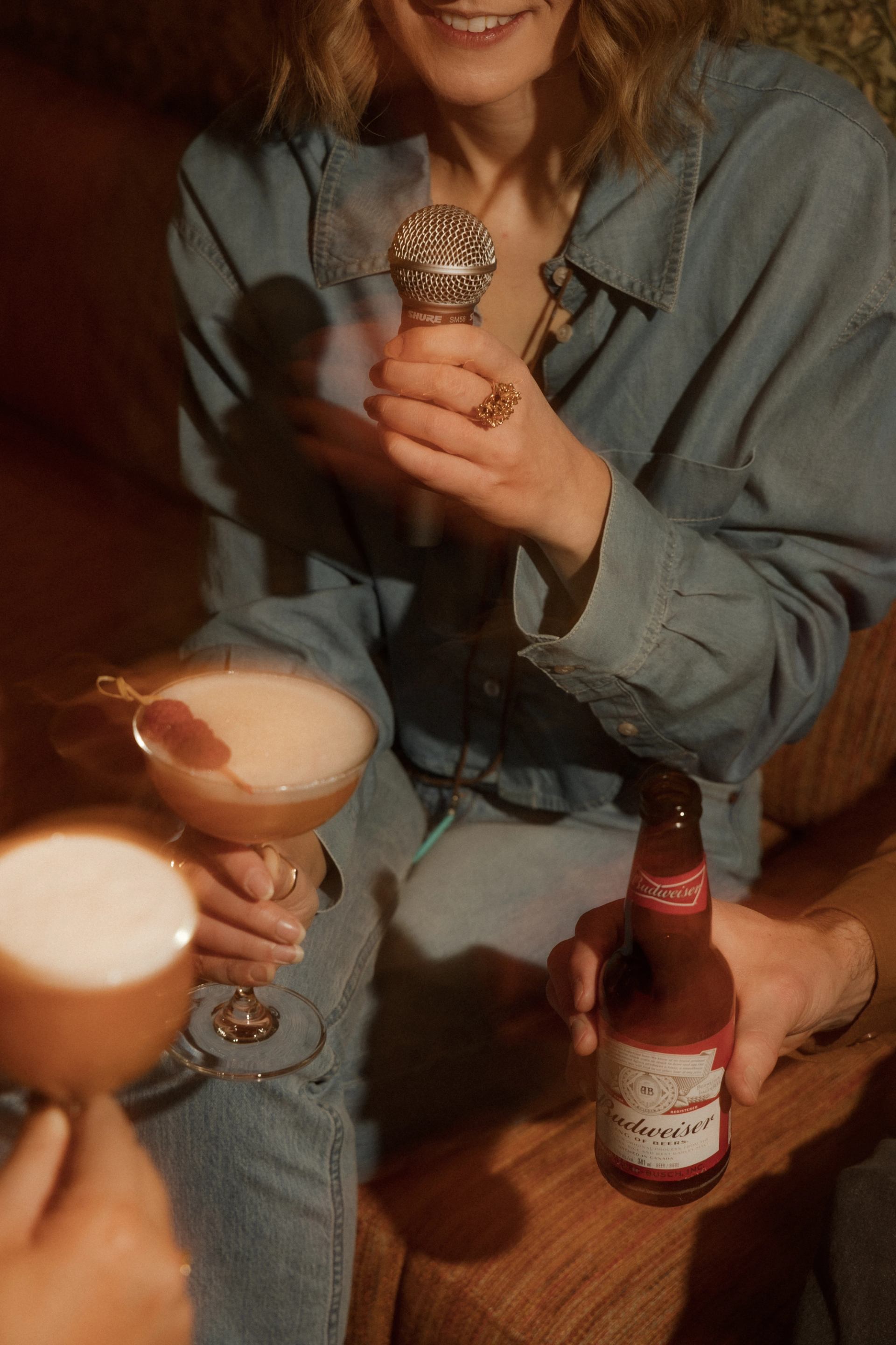 Person holding a microphone while others raise cocktails and a beer in a cozy bar setting.