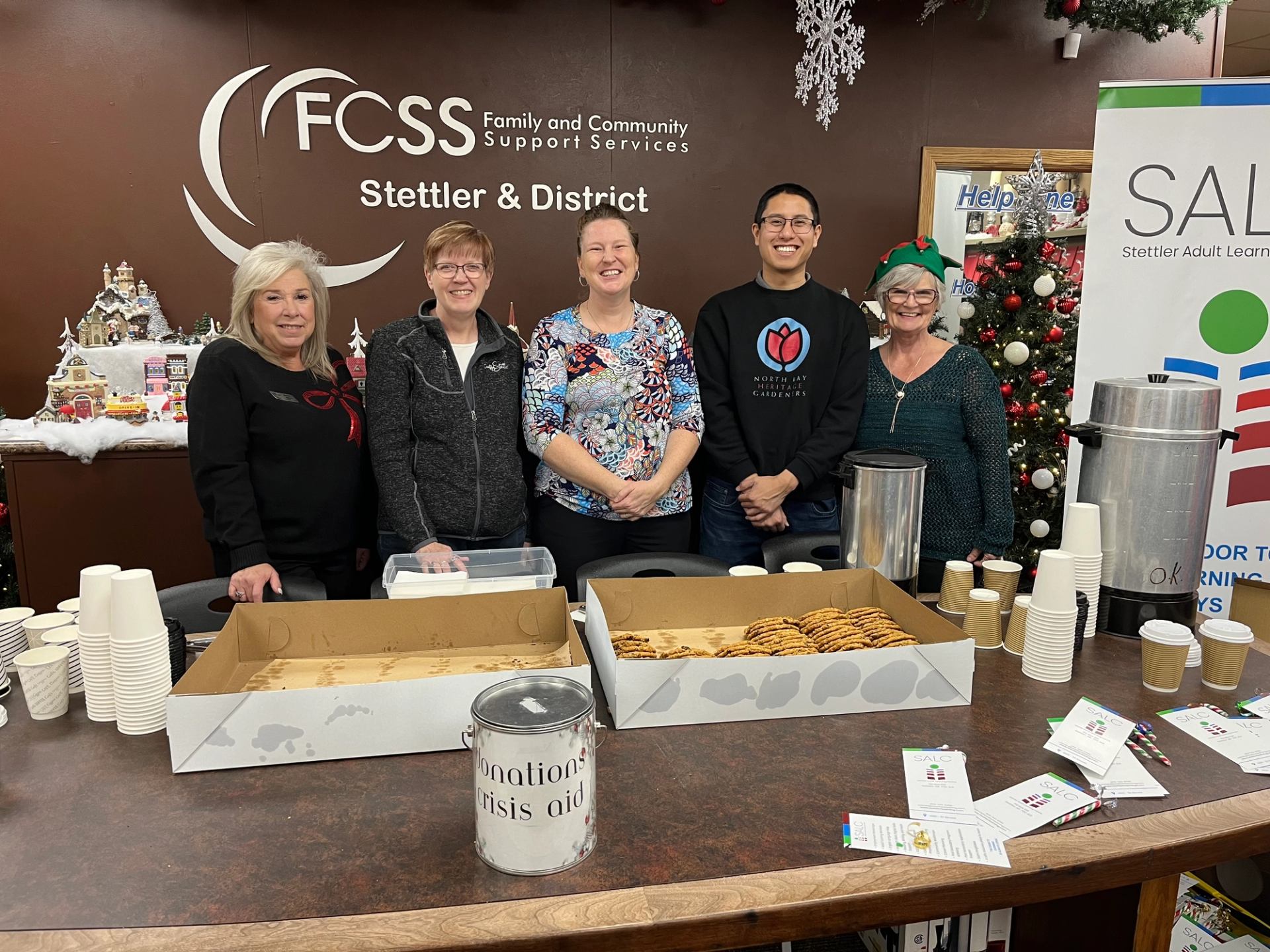 People stand behind table with cookies, donations box, and festive decor.