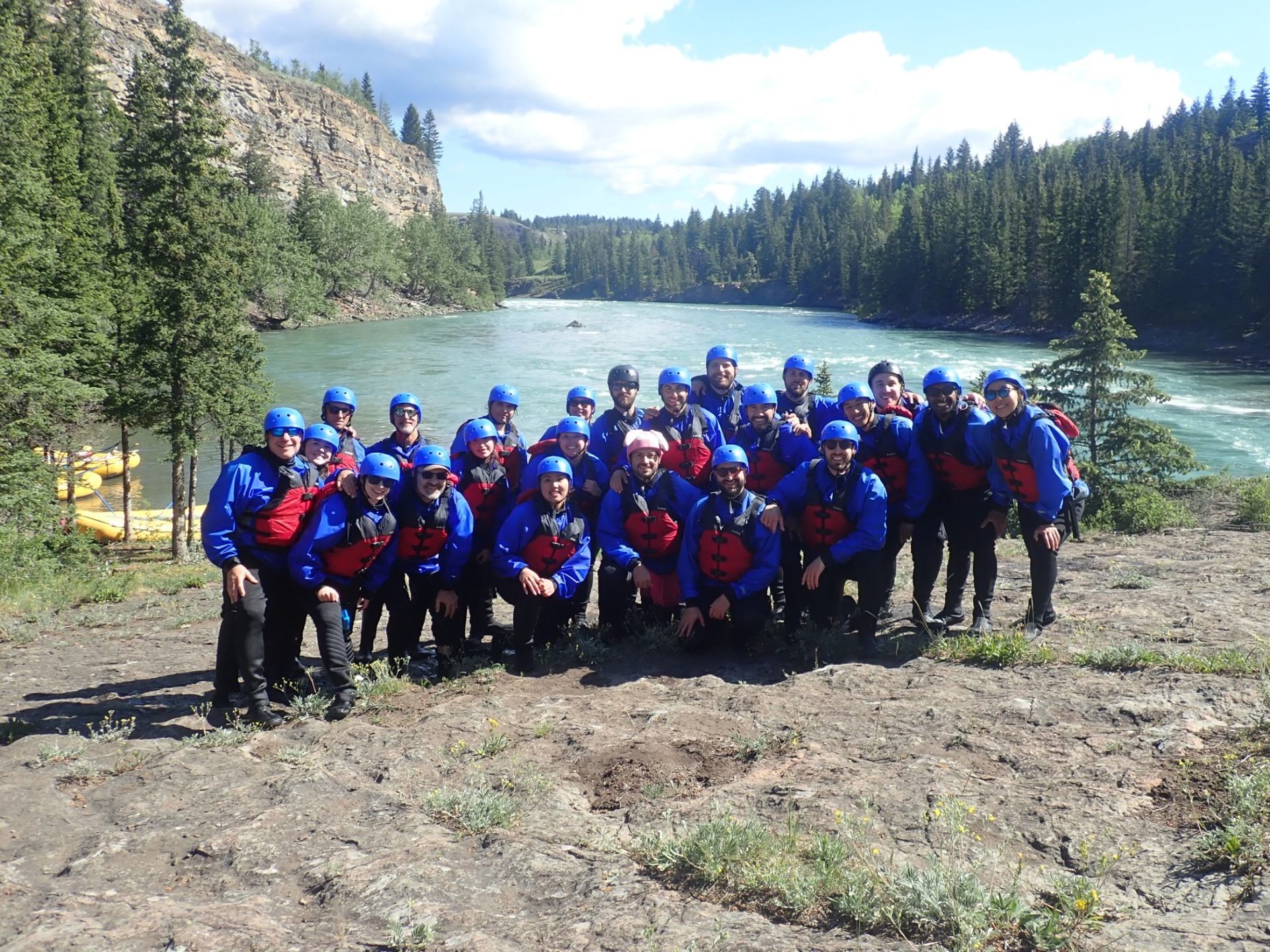 Group in blue rafting gear posing on rocky riverbank beside forested canyon river.