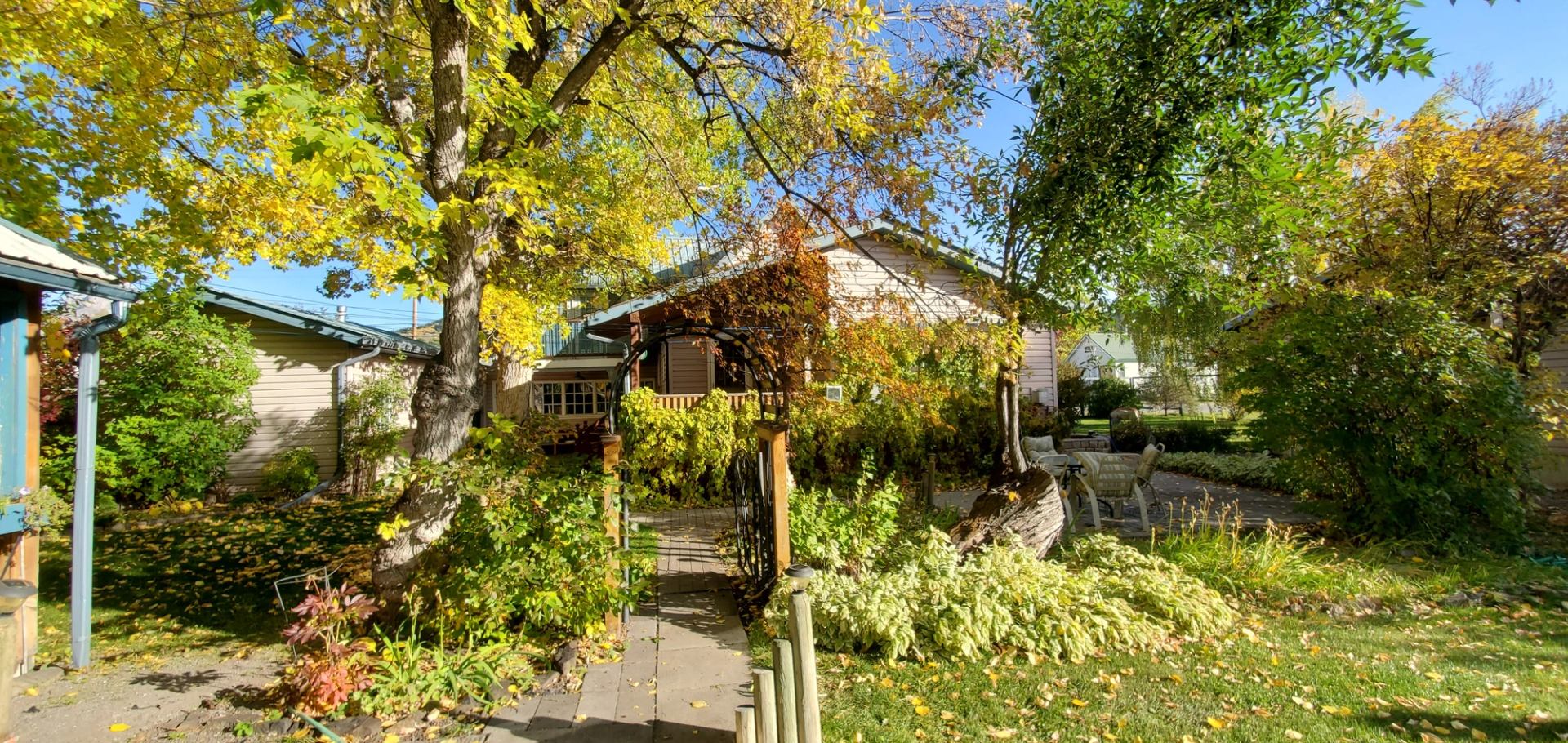 Garden path through trees with yellow leaves leading to cottage porch and patio chairs.