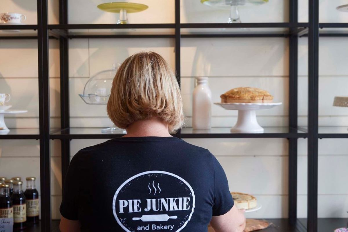 Person wearing a Pie Junkie and Bakery shirt standing in front of shelves displaying pies and cake stands.