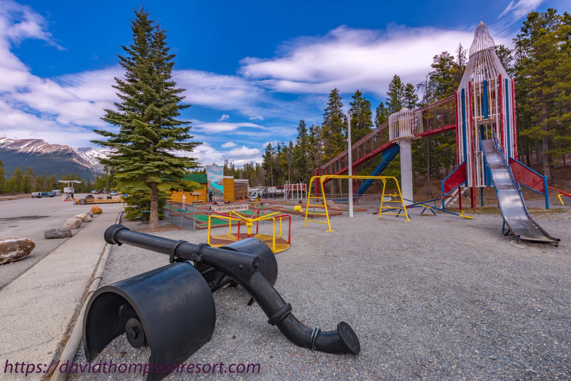 Playground with slides, swings and climbing structures beside evergreen trees and mountains.