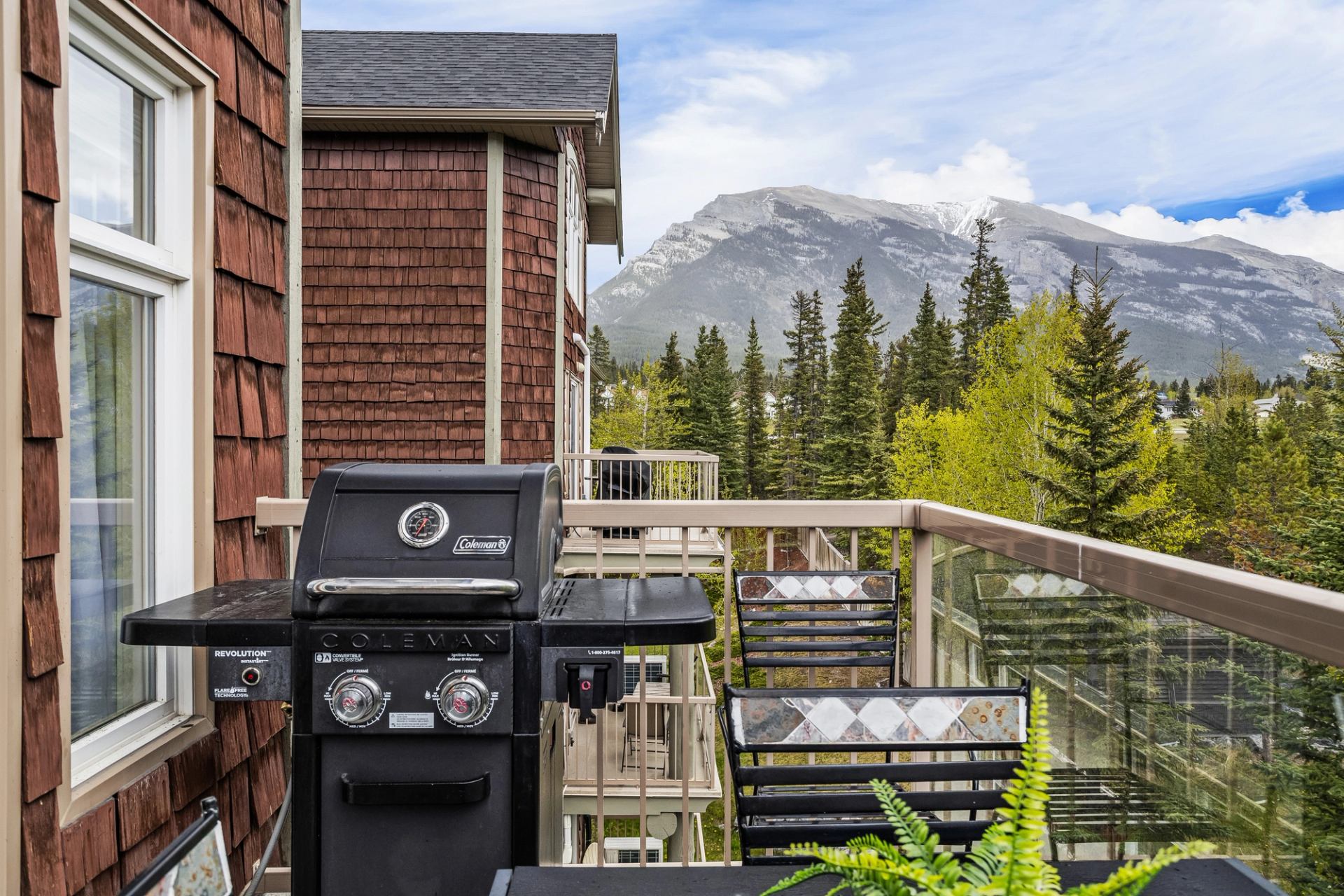 Balcony with barbecue and chairs overlooks evergreen trees and a rocky mountain.
