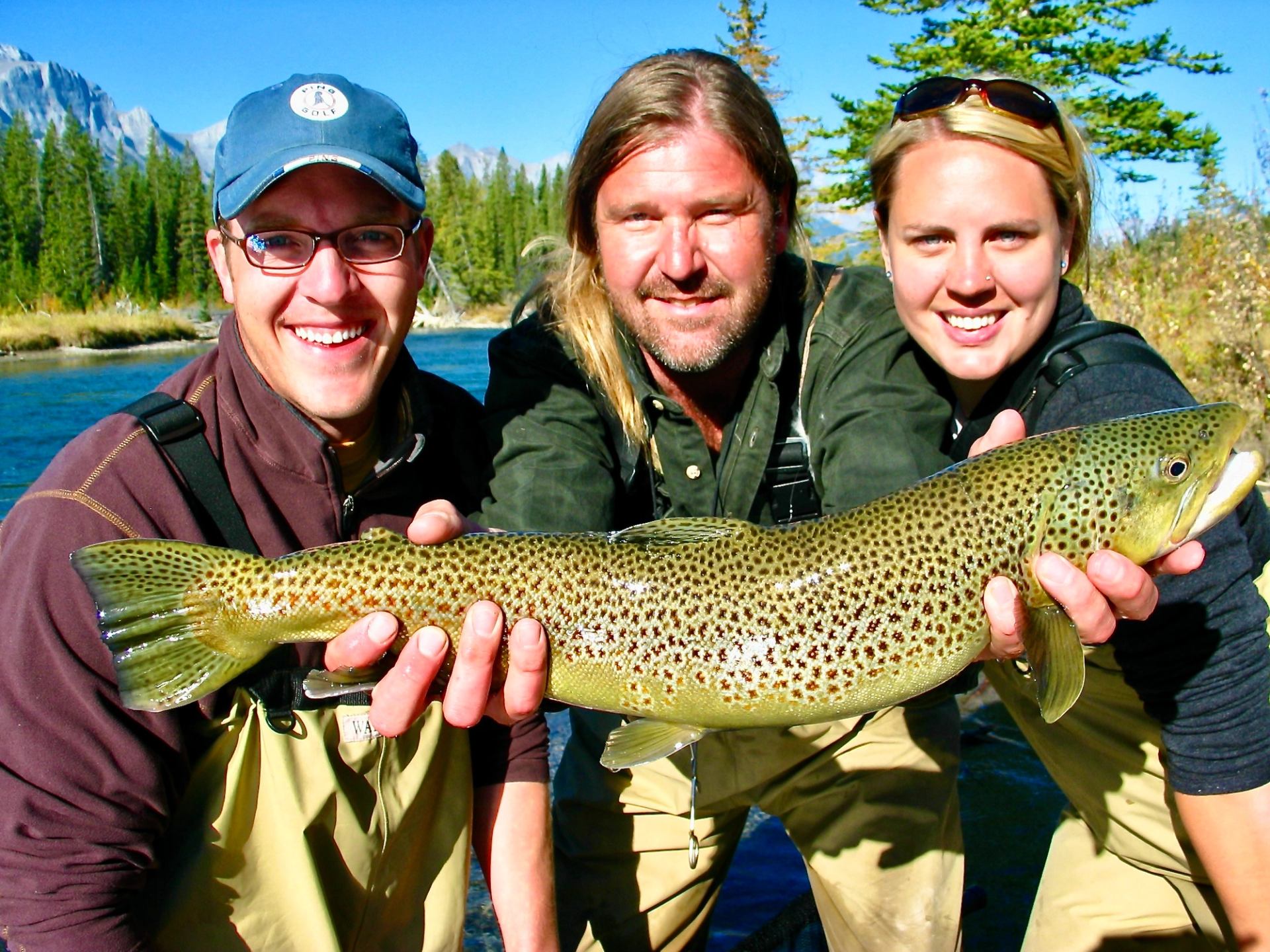 Three people in waders hold a large speckled fish beside a forested river.