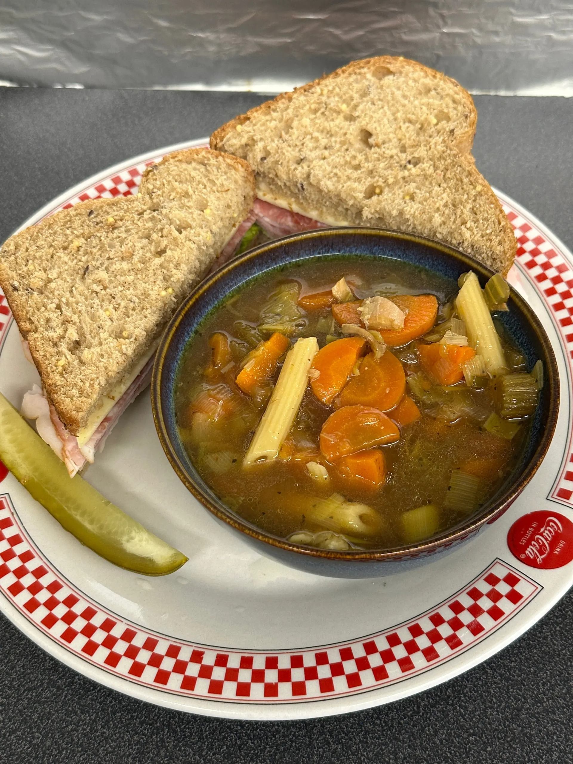 Bowl of vegetable pasta soup with sandwich halves and pickle on checkered plate.