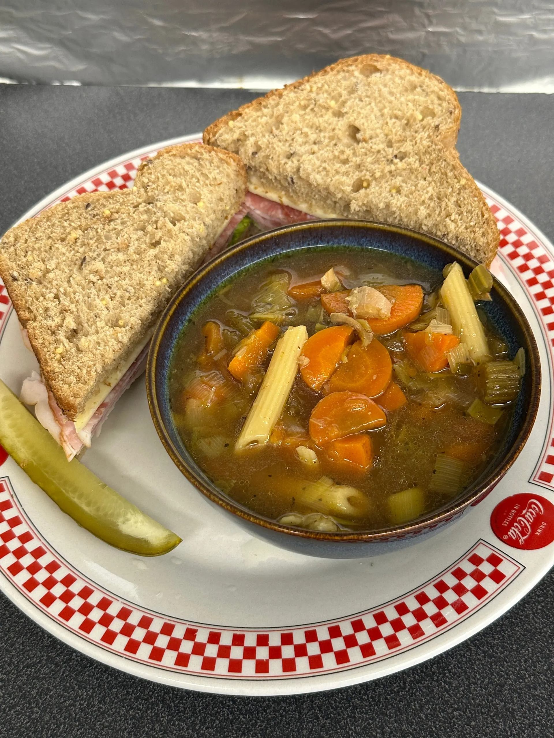 Bowl of vegetable pasta soup with sandwich halves and pickle on checkered plate.