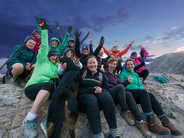 Group of hikers on a rocky summit at sunset, smiling with a dog and dramatic sky in the background.