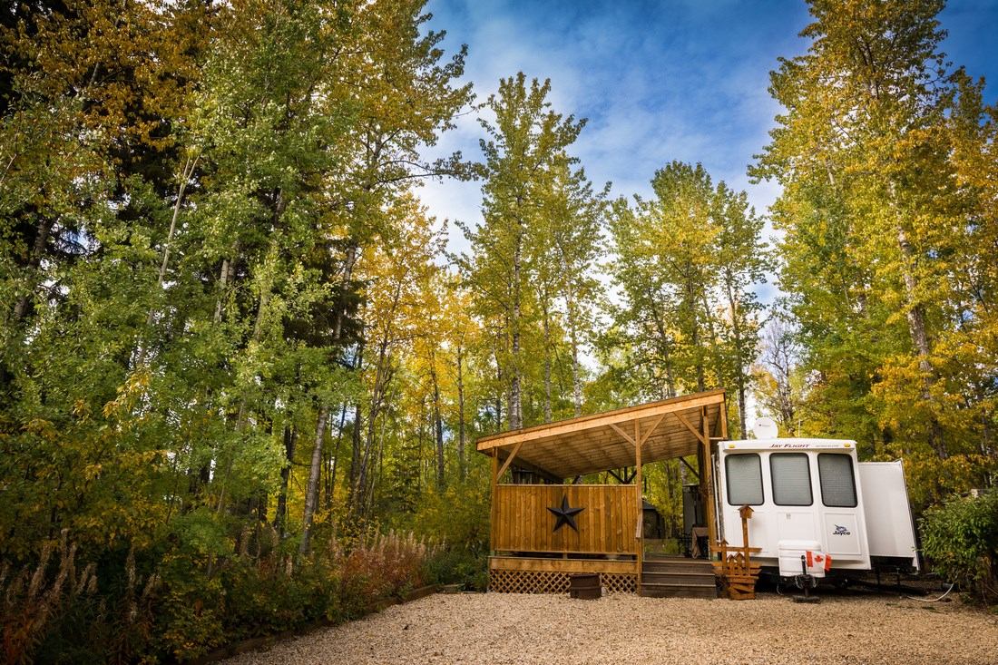 Wooden deck with star beside white trailer, surrounded by tall green and yellow trees.