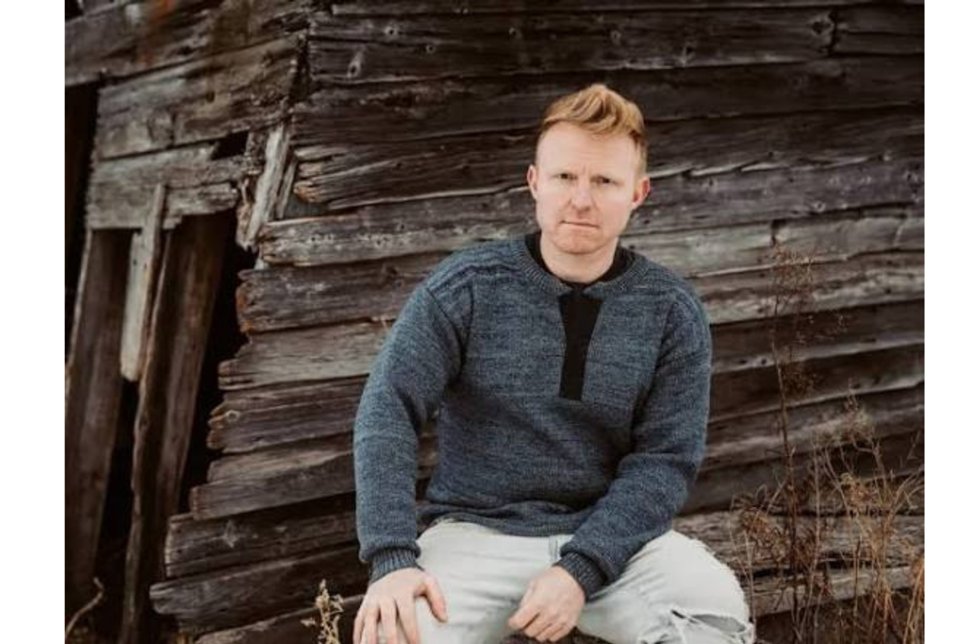 Person in a grey sweater sits against a weathered wooden wall.