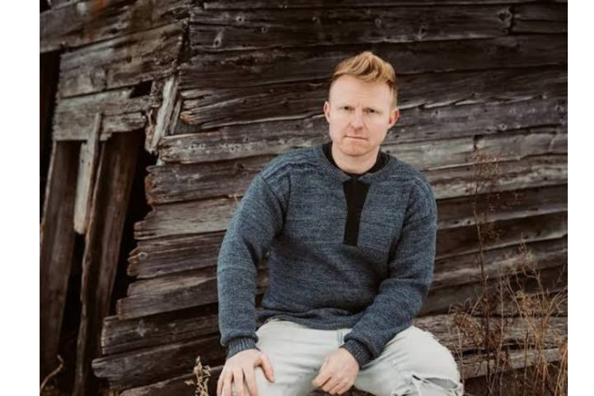 Person in a grey sweater sits against a weathered wooden wall.