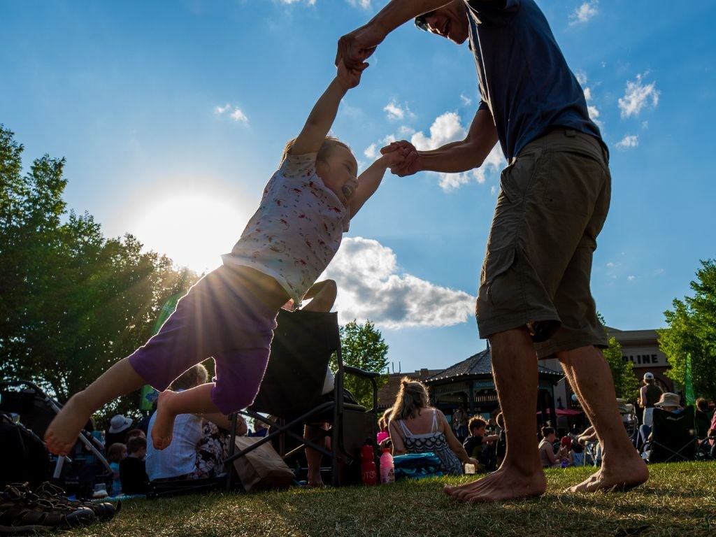 Child being swung by adult at sunny plaza event.