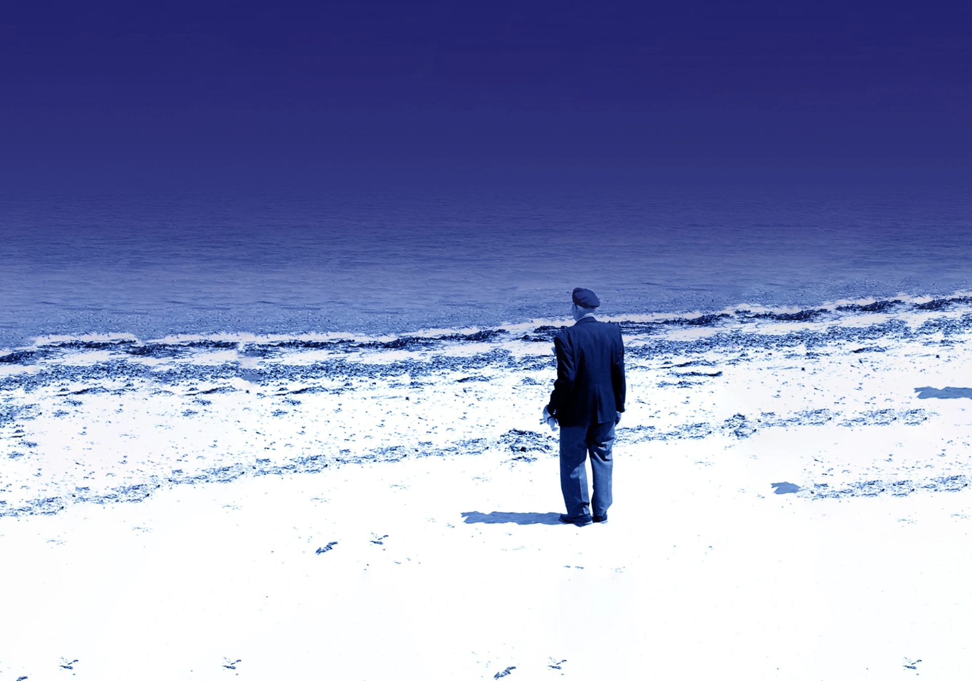 Person in dark clothing stands on bright beach facing calm blue ocean horizon.