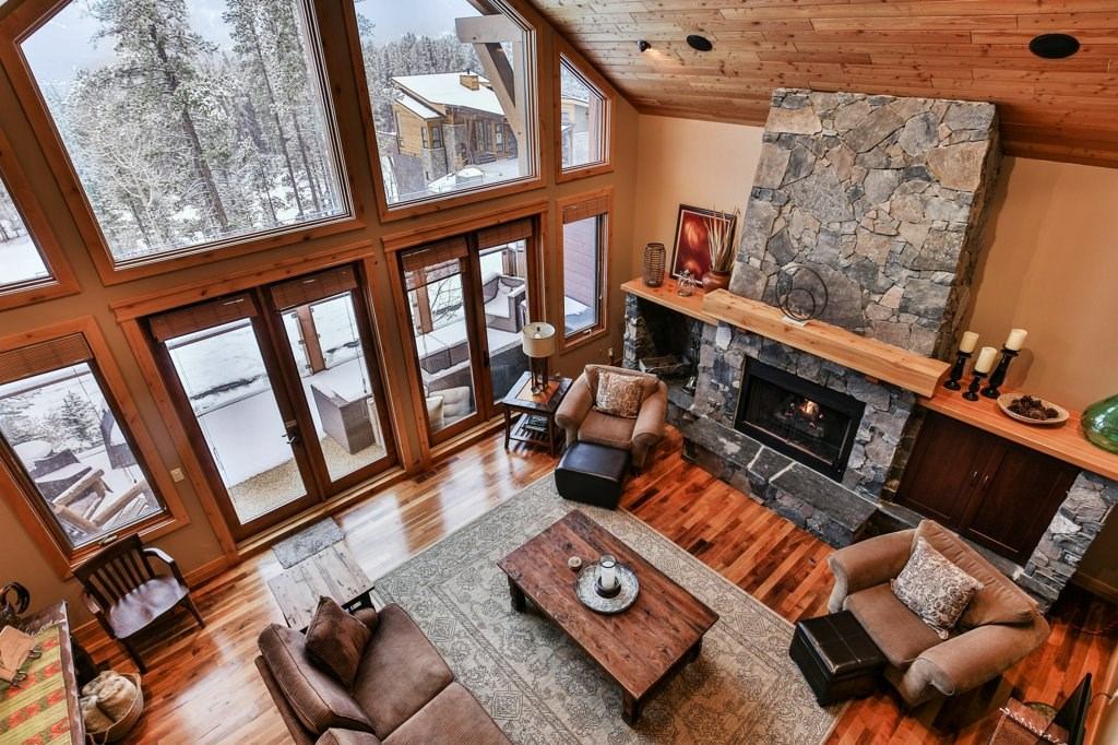 Rustic living room with stone fireplace, wood floors, and large windows overlooking snowy trees.