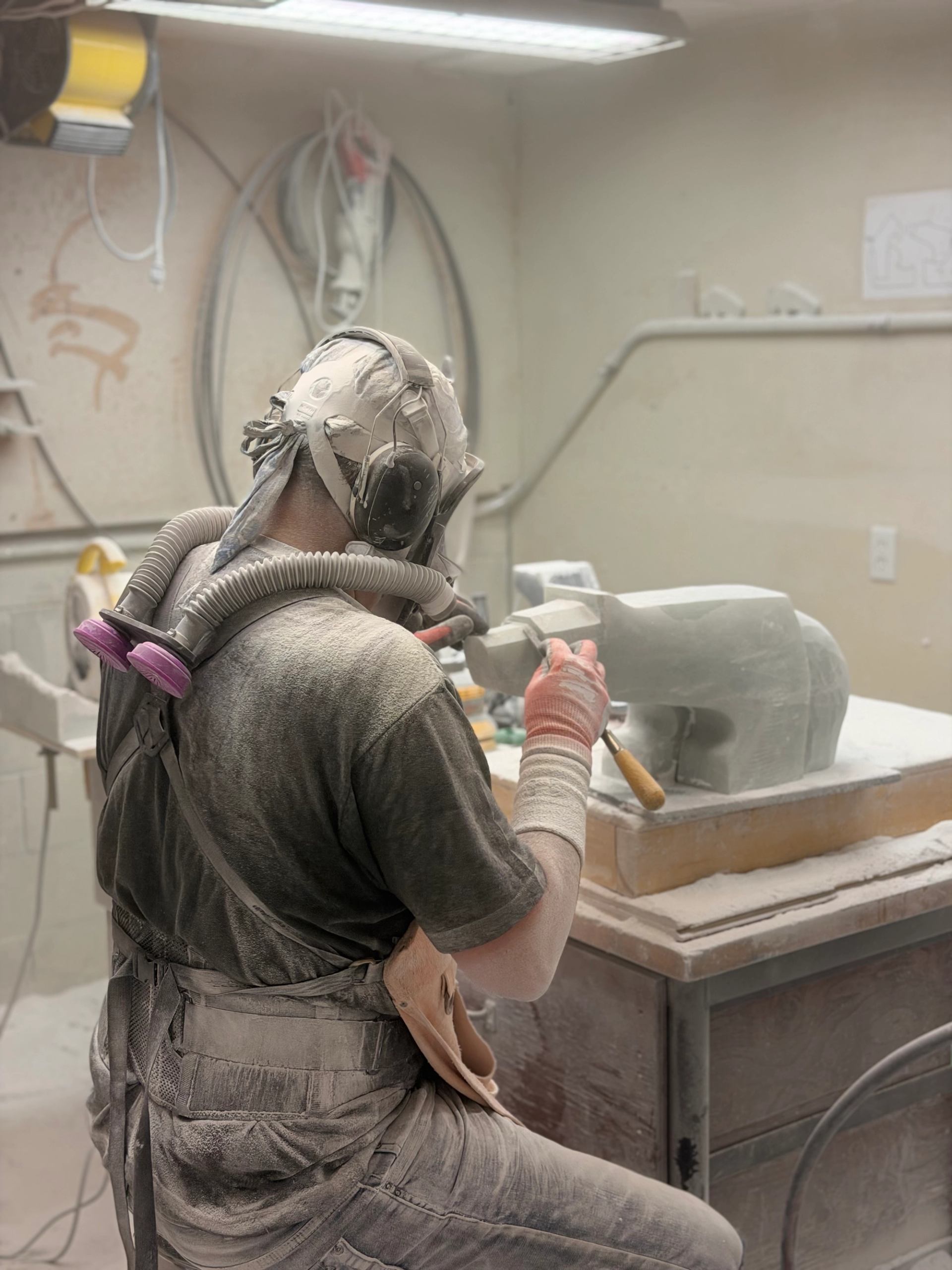 Stone carver wearing respirator and ear protection shapes sculpture in dusty workshop.