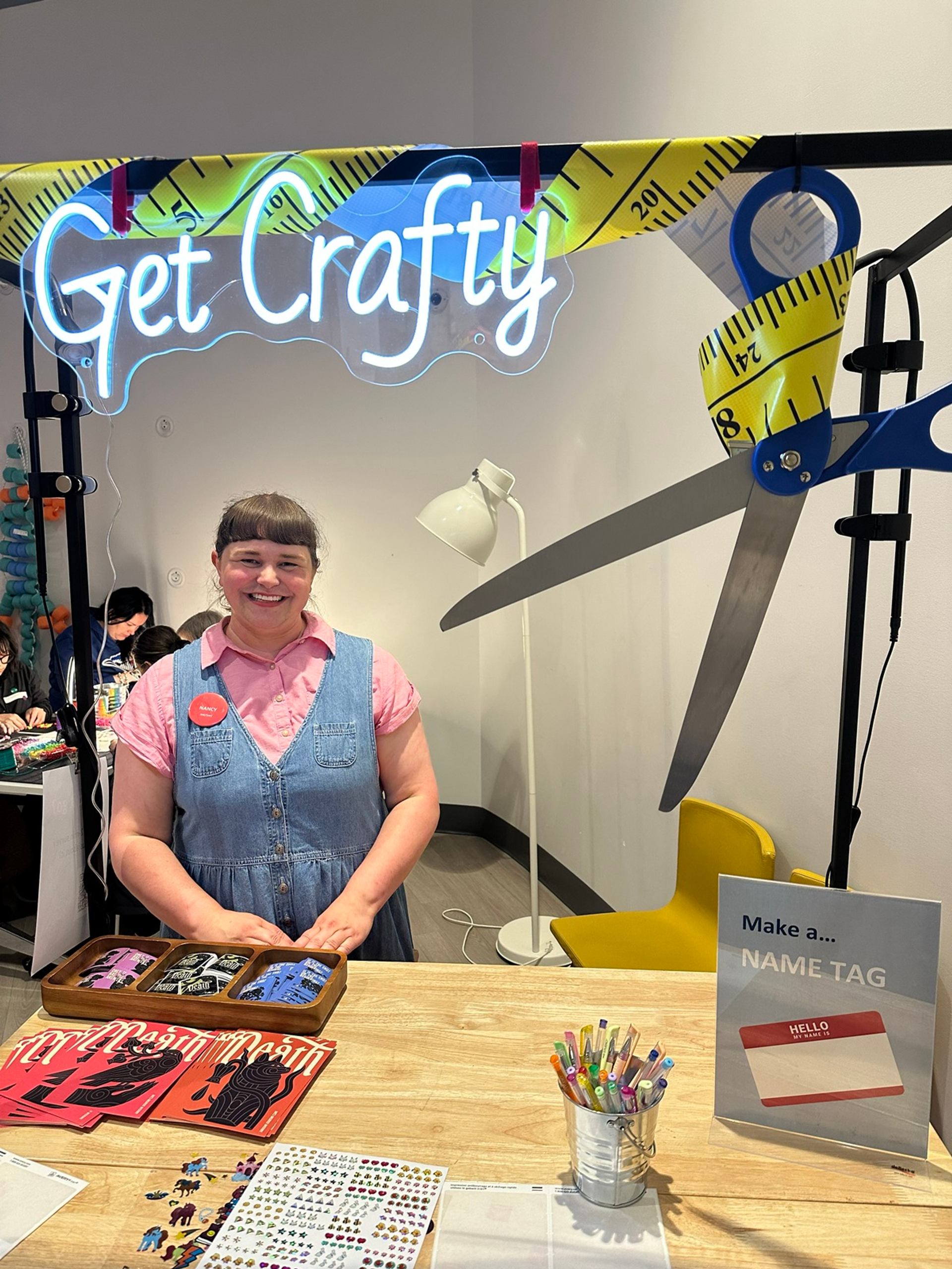 Person stands at a name tag craft table beneath a glowing Get Crafty sign.