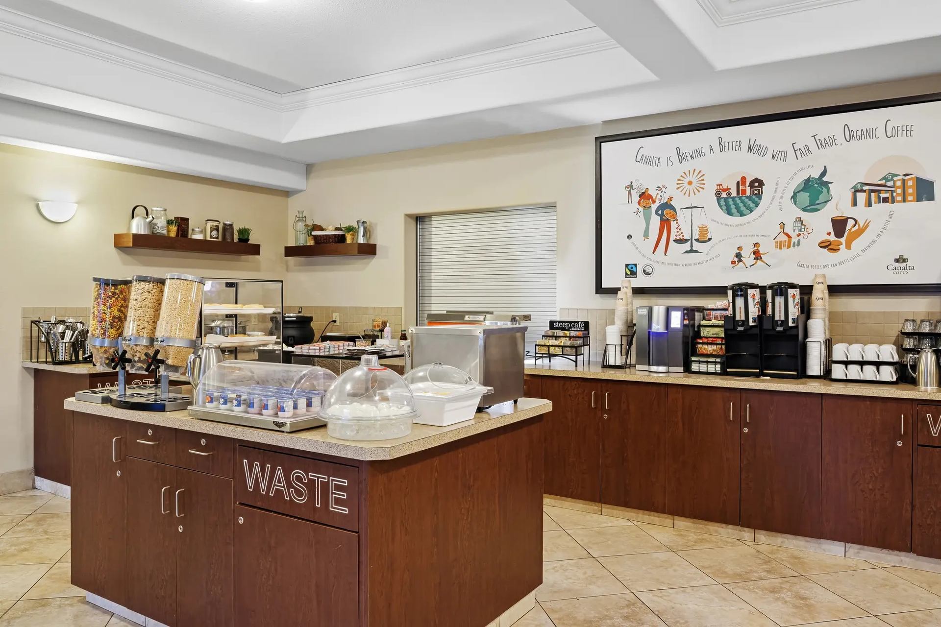 Hotel breakfast buffet area with cereal dispensers, covered pastries and coffee station.