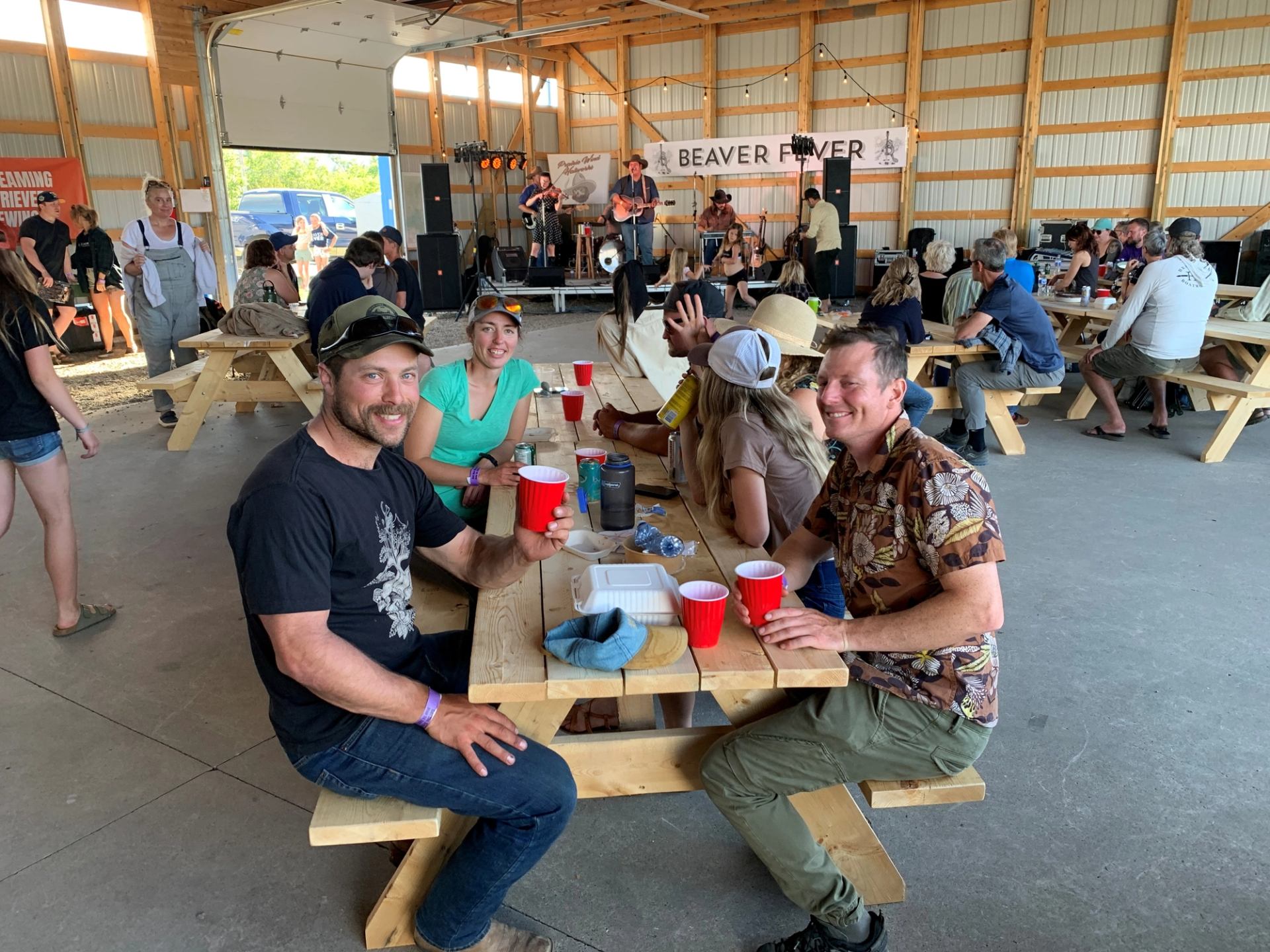 People gather at picnic tables in a large event hall, with a band performing under a "BEAVER FEVER" banner. Two men in the foreground smile at the camera, holding red cups.