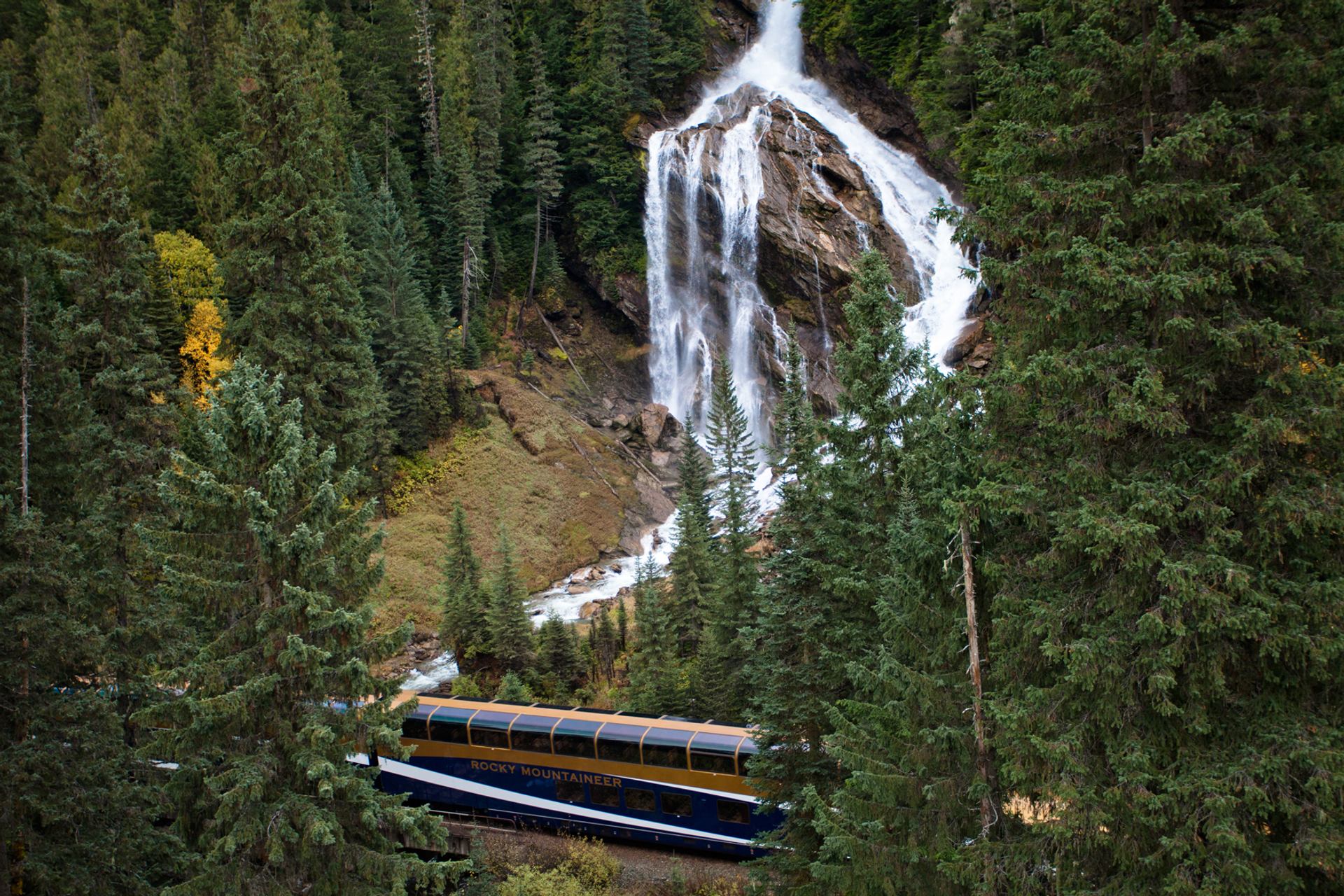 Rocky Mountaineer train passing waterfall and forested cliffs.