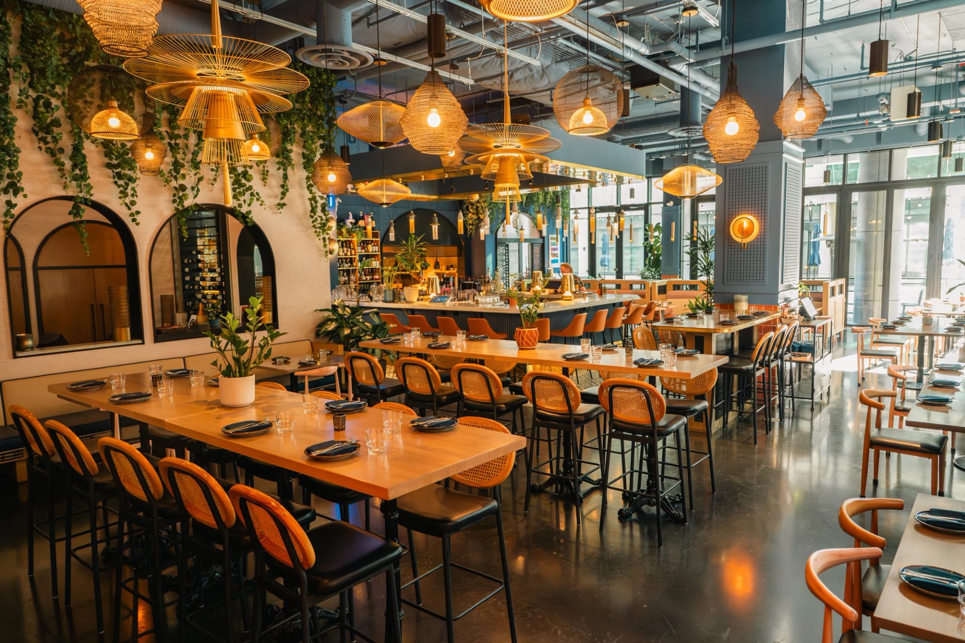 Modern restaurant interior with hanging woven lights, long wooden tables and central bar.