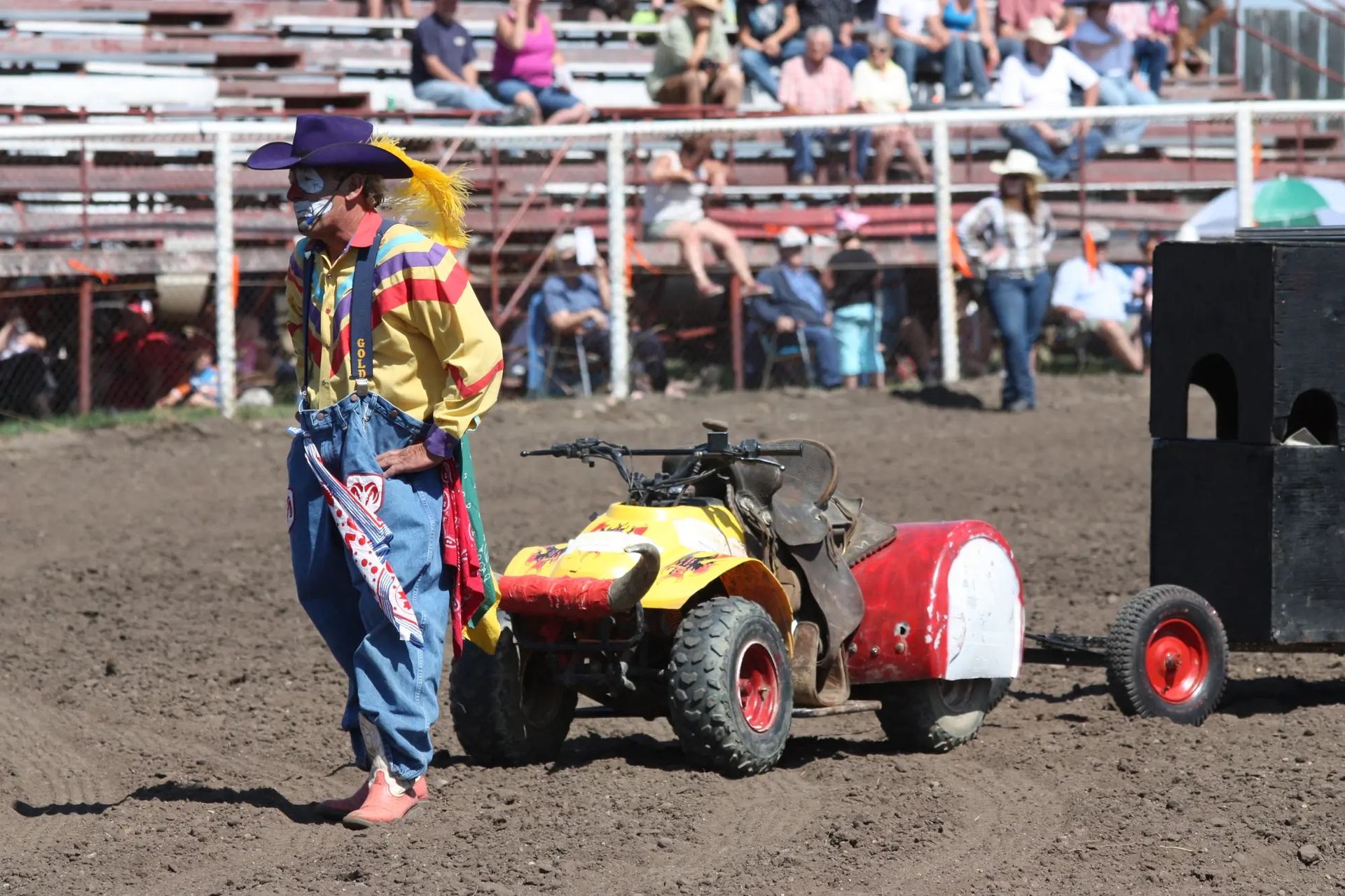 Rodeo clown in colorful outfit standing beside ATV in arena.