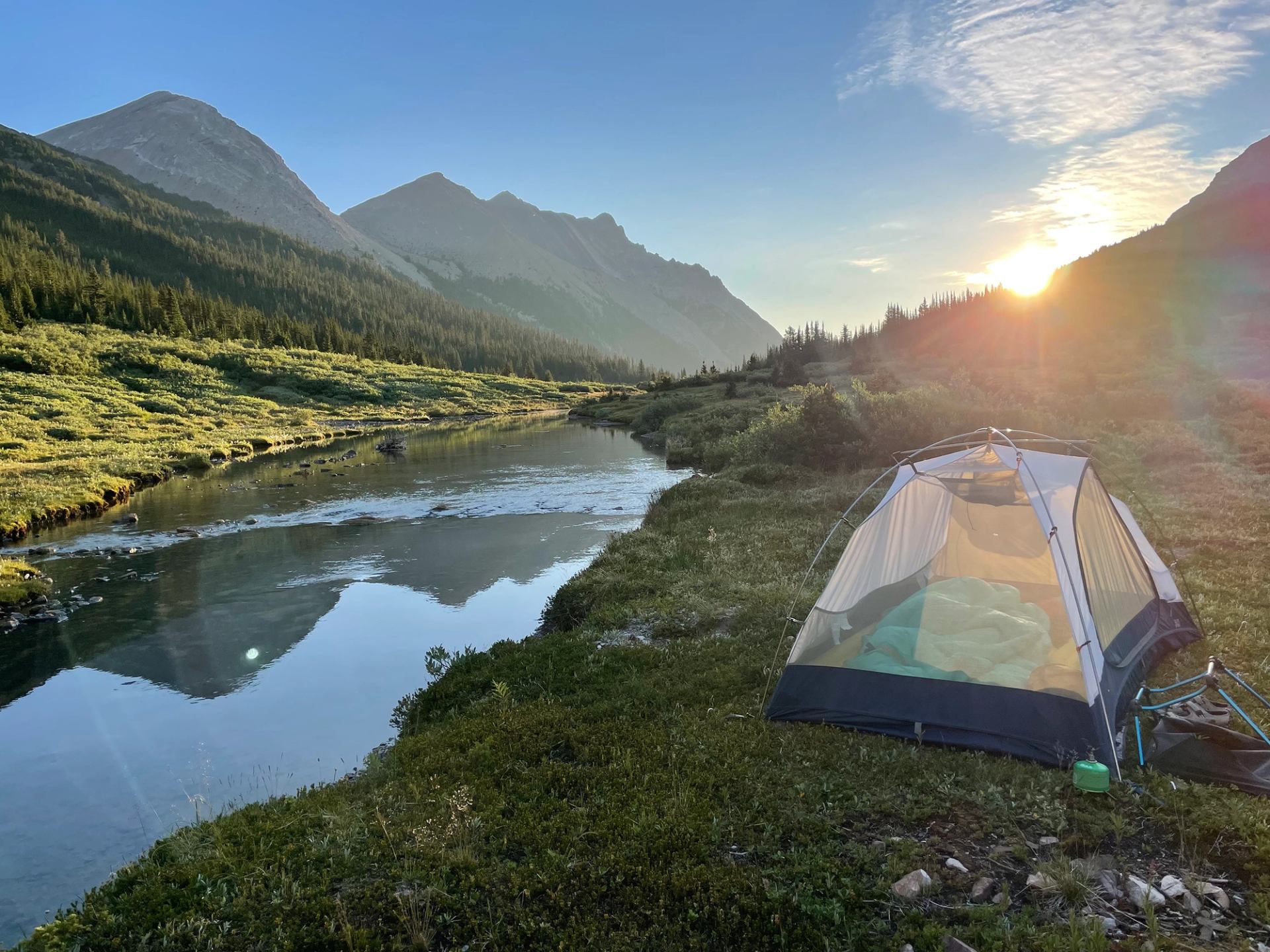 Tent beside calm river with mountains and trees under low sun in clear sky.