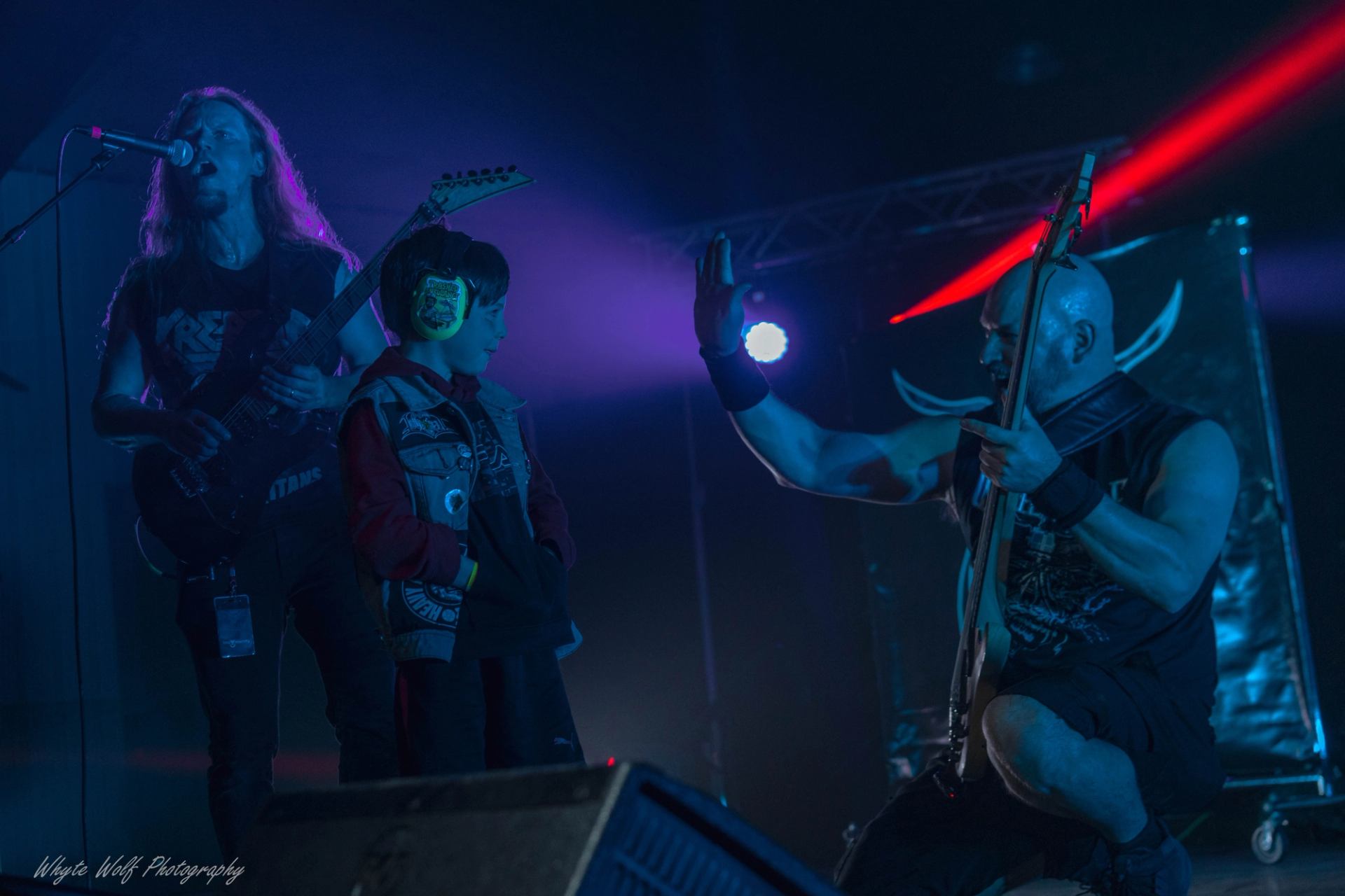 Decimate Metalfest shows musicians on stage as a young fan wearing ear protection reaches toward a guitarist under colored lights.