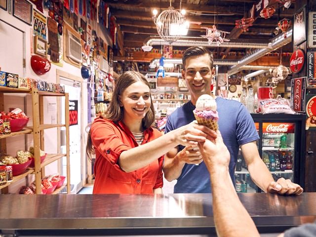 Two people hold ice cream cones inside a candy shop lined with shelves.
