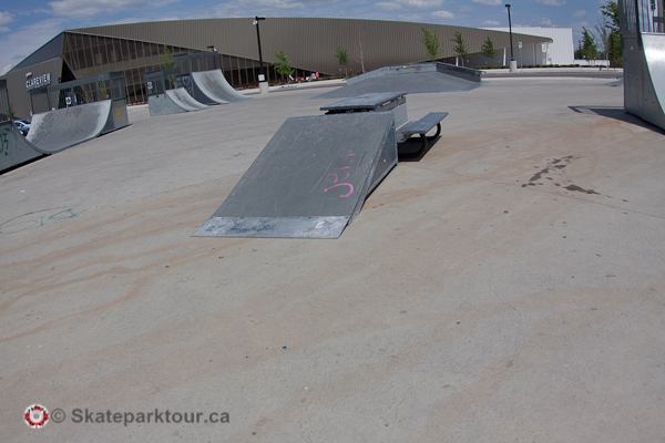 Metal funbox ramp and skate feature positioned on a broad concrete surface at an outdoor park.